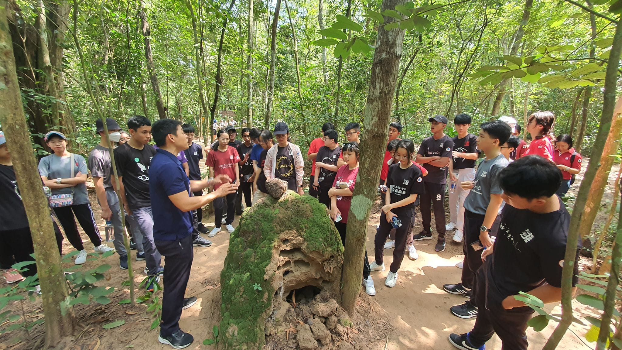 A guide speaks to a large group of young people gathered around a moss-covered rock formation in a forest.