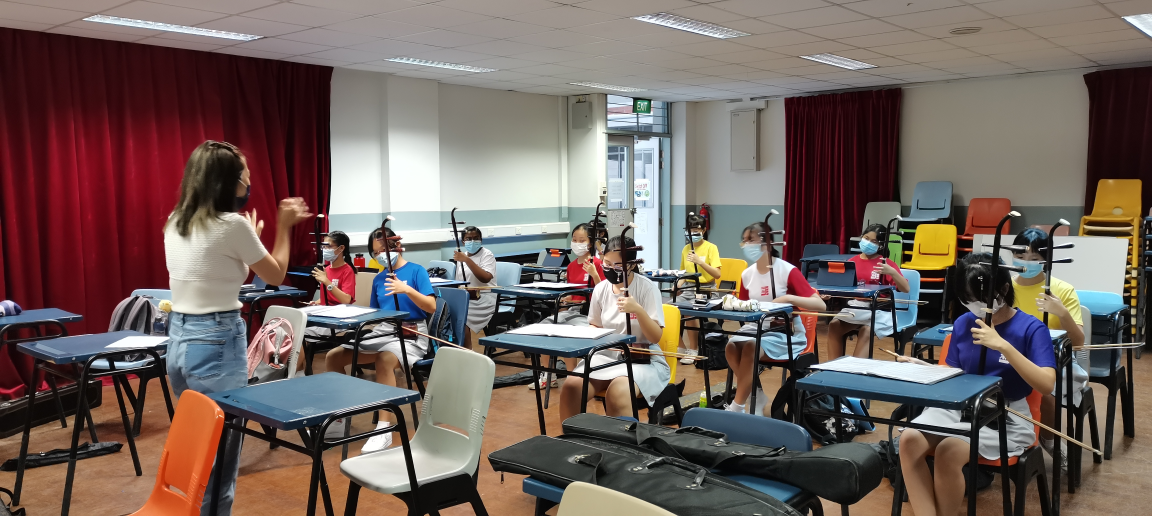 Students wearing masks sit at desks holding string instruments while a teacher stands and gestures at the front of a classroom.