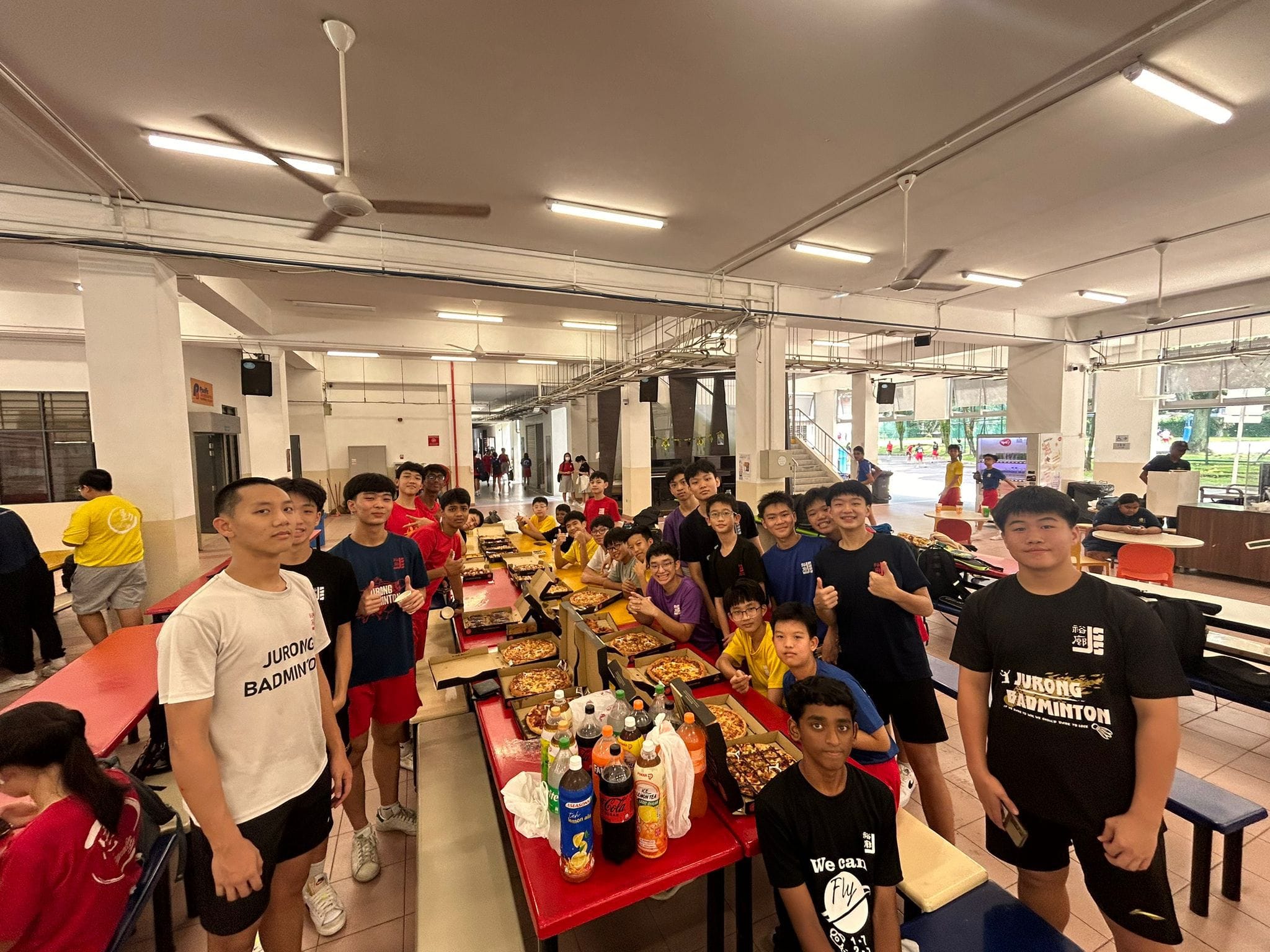 A large group of teenage boys in Jurong Badminton shirts pose around tables with open pizza boxes and drinks.
