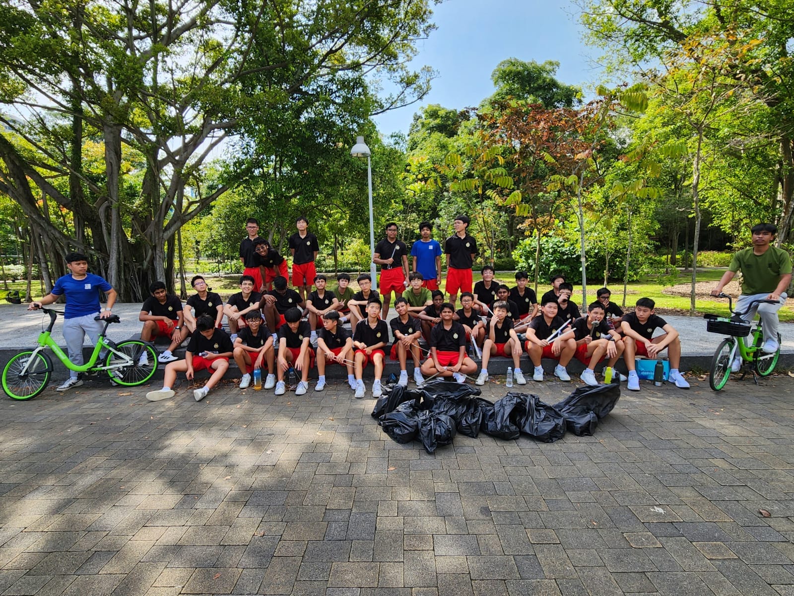 A large group of students in black and red uniforms pose in a park with black trash bags and bicycles nearby.