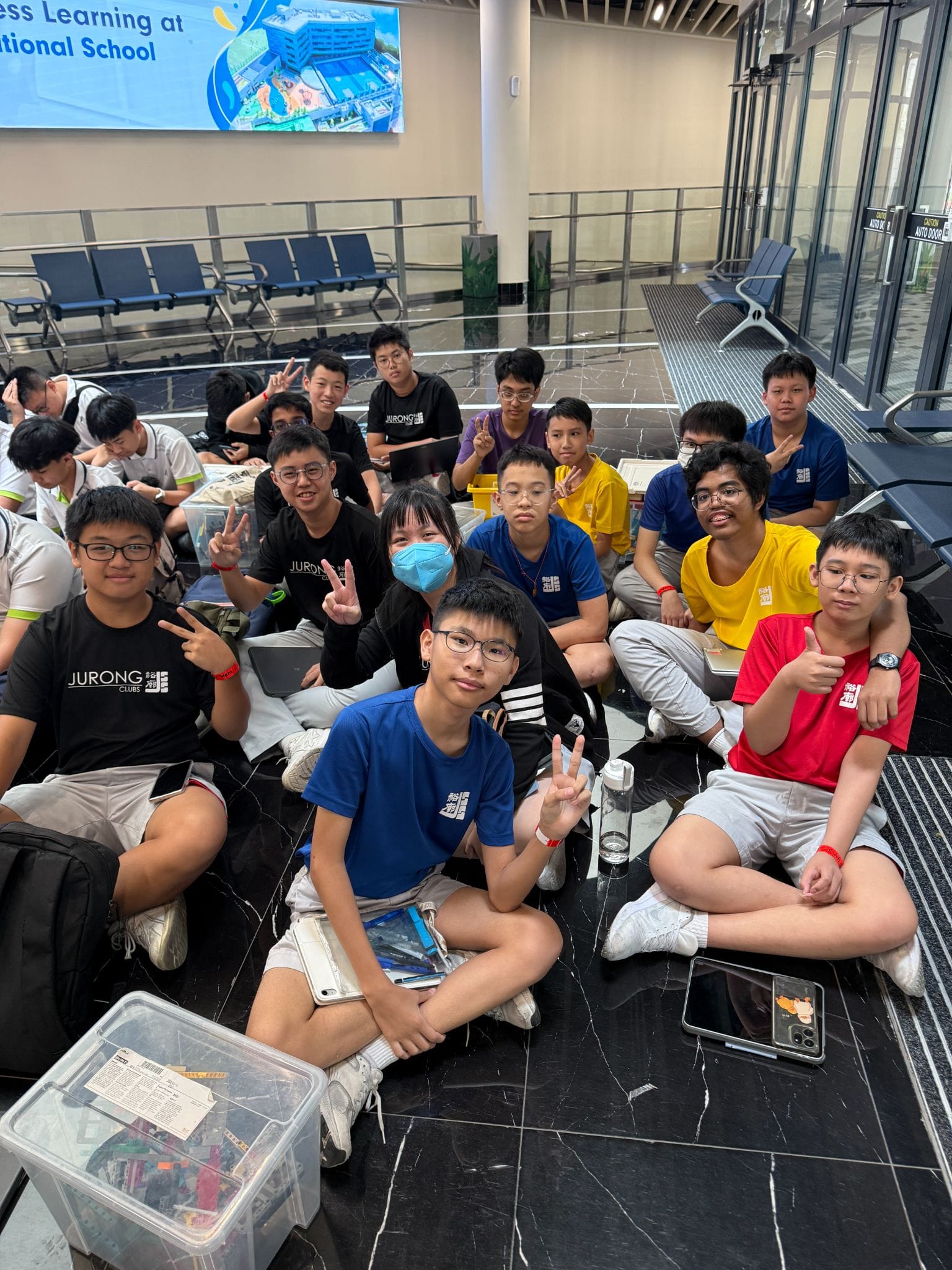 Group of students in Jurong school shirts sitting on a floor, smiling and making peace signs at the camera.