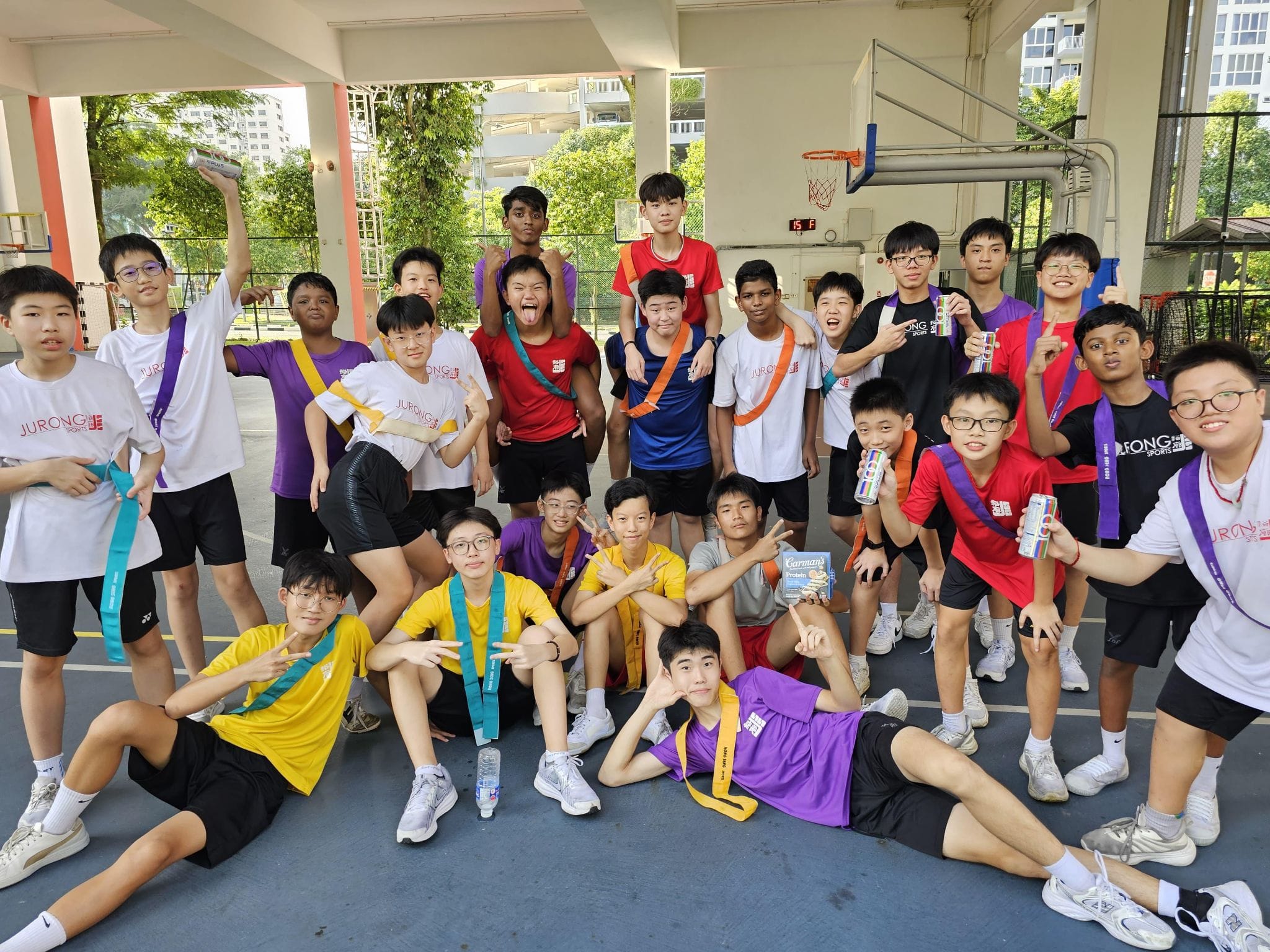 A large group of teenage boys in colorful sports jerseys pose and smile on an outdoor basketball court.