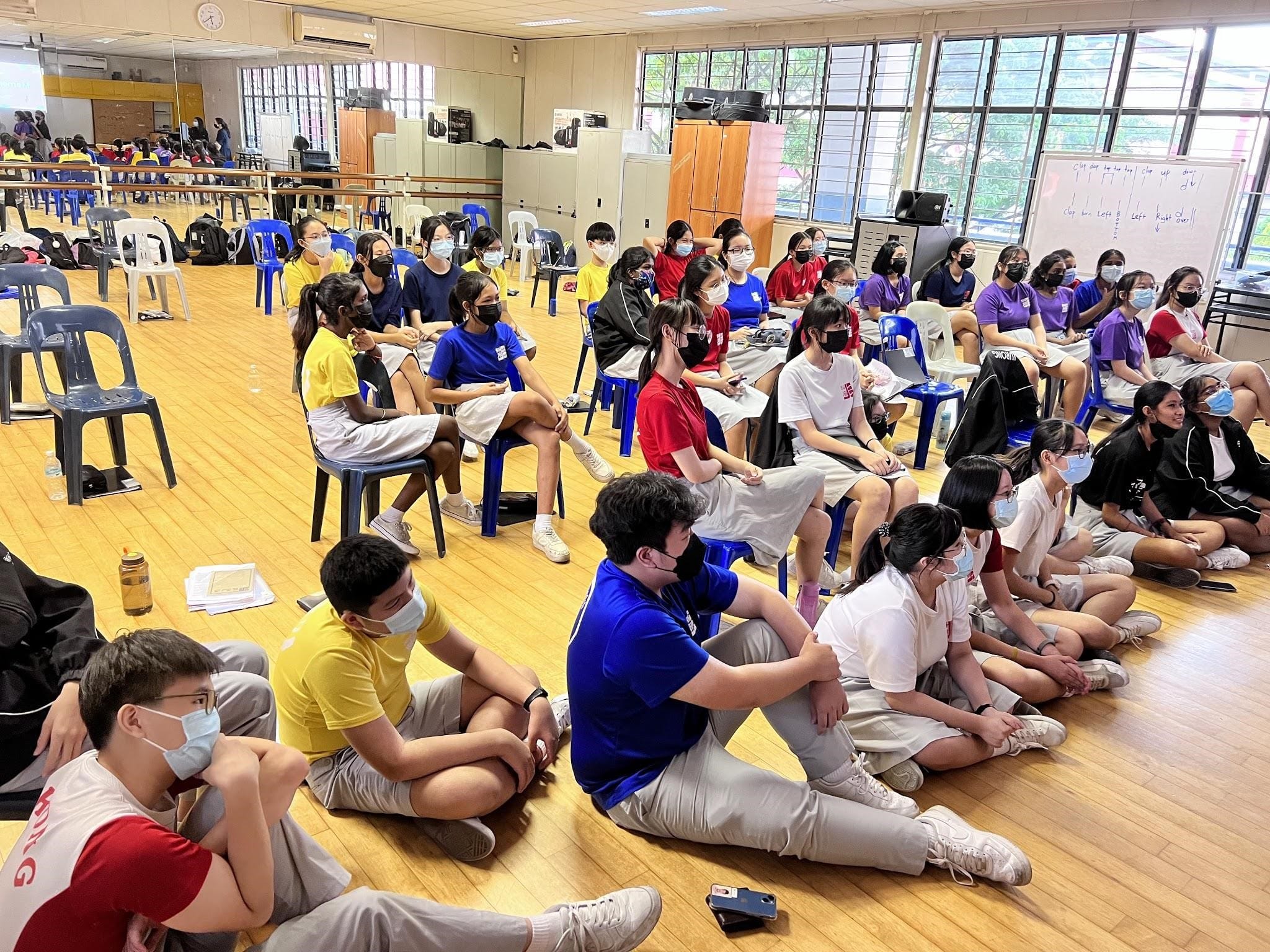 Students wearing face masks sit on the floor and chairs in a school hall during a group session.
