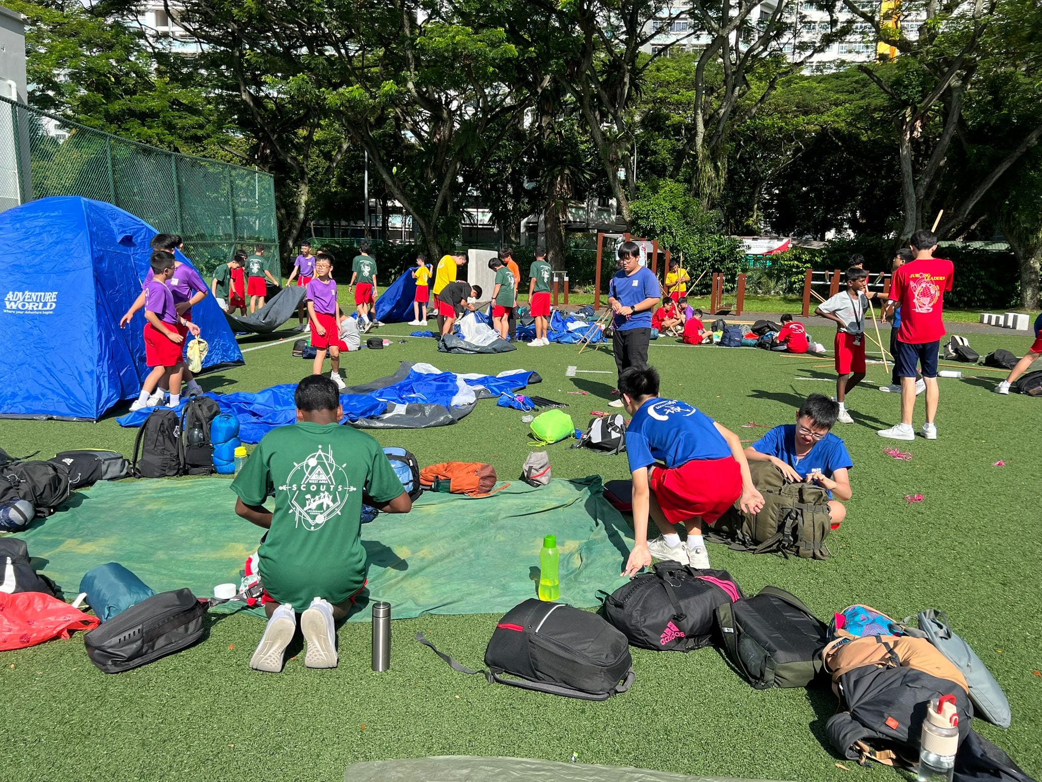 Youth scouts set up tents and sort gear on a green field surrounded by trees during an outdoor camp activity.