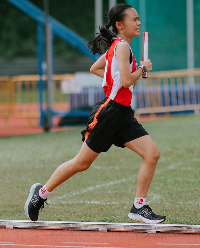 A young female runner in a red and white jersey sprints on a track holding a red relay baton.