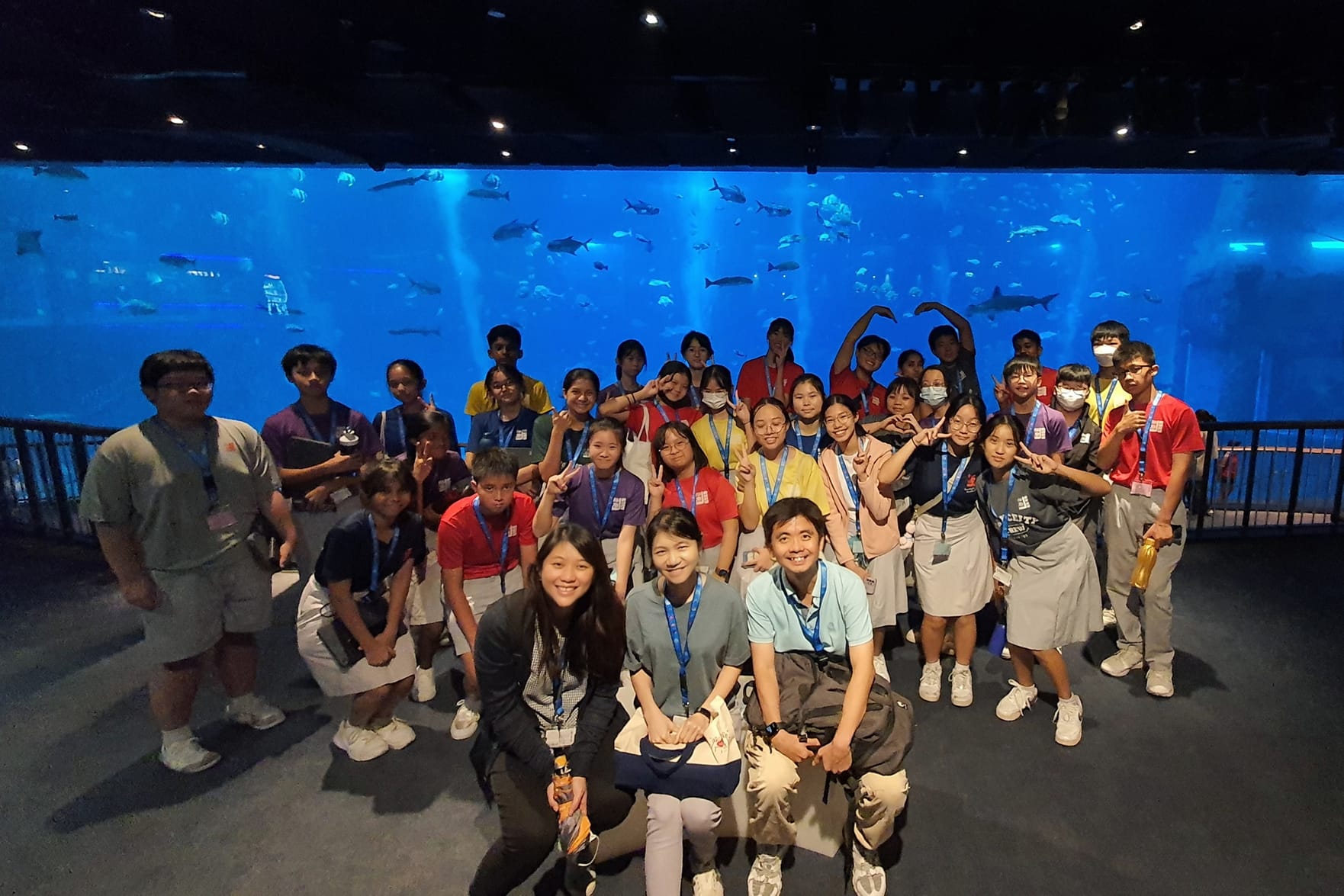 A large group of students and adults pose in front of a giant blue aquarium tank filled with fish.