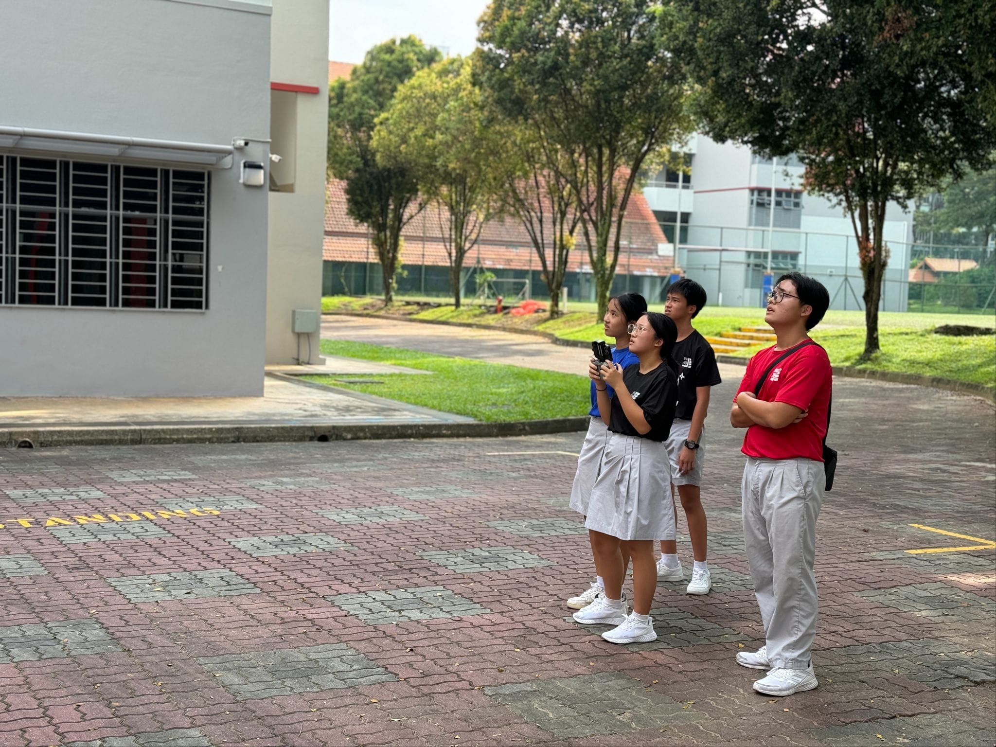 Four students in school uniforms stand on a brick courtyard, looking upward near a gray building with trees behind.