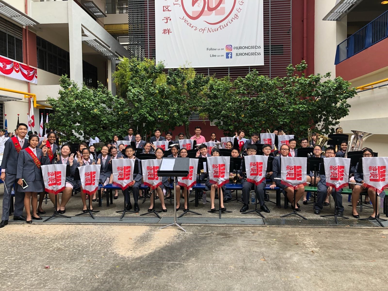 A school band in grey uniforms poses outdoors at Jurong Secondary School's 55th anniversary celebration.