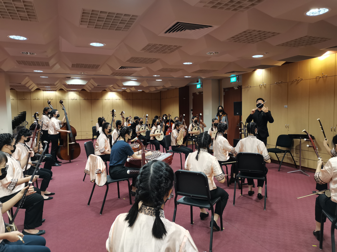 A conductor leads a Chinese orchestra ensemble of masked musicians in pink uniforms rehearsing in an indoor room.