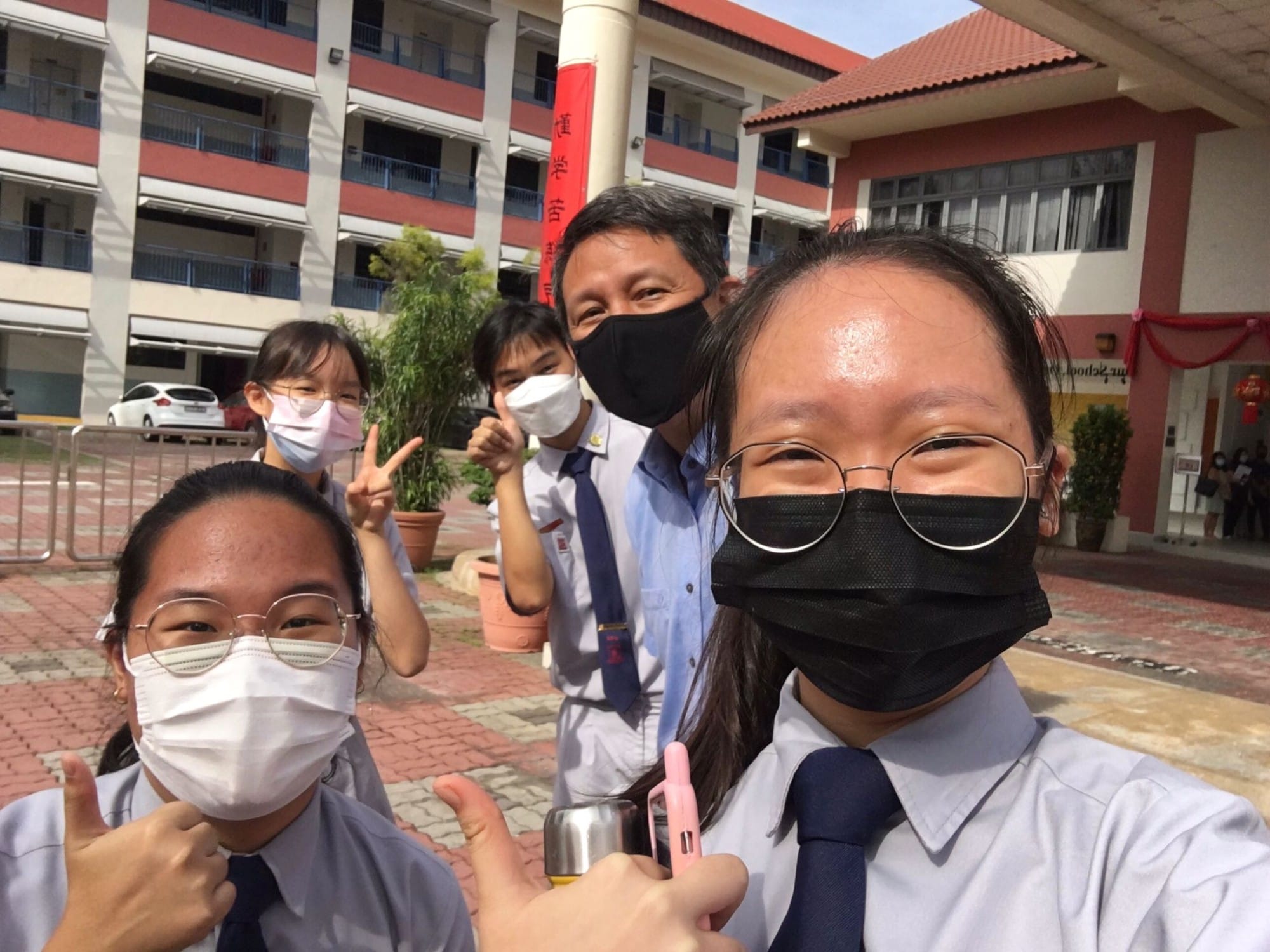 Four people wearing face masks pose for a selfie outdoors in front of a multi-story school building.