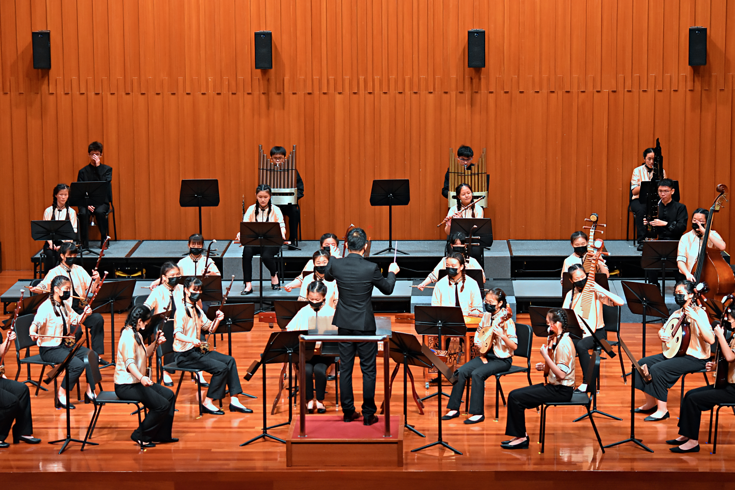 A conductor leads a youth Chinese orchestra on a wooden stage, with masked musicians playing traditional instruments.