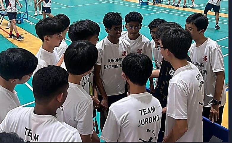 A group of teenage boys in white Team Jurong Badminton shirts huddle together on an indoor court.
