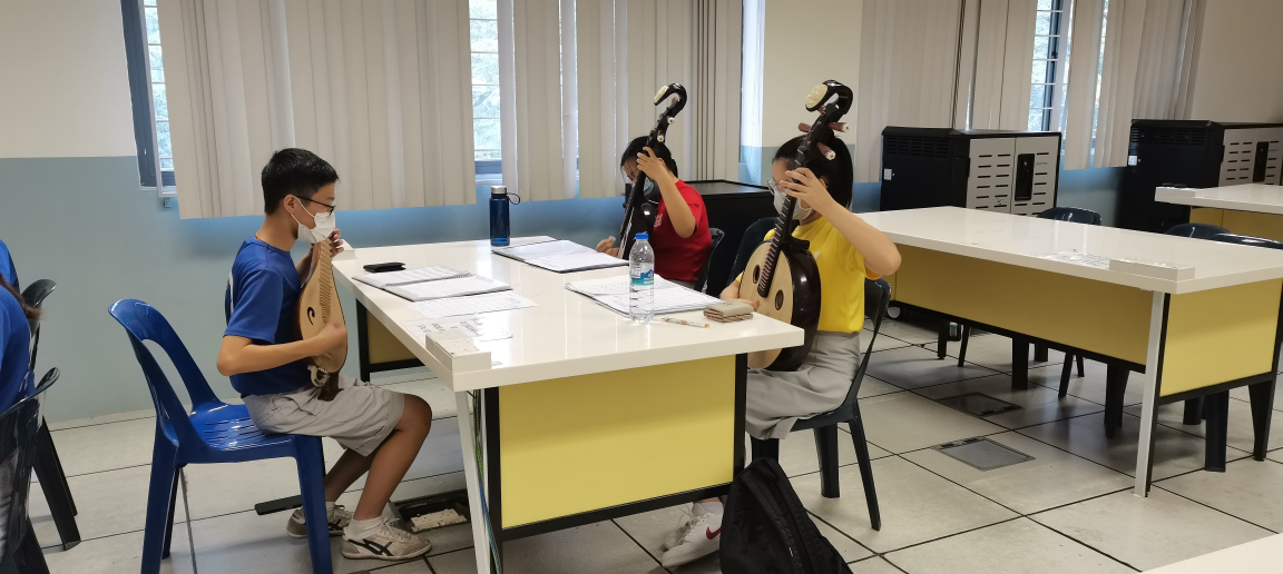 Three masked students sit at a table in a classroom playing traditional Chinese lute instruments called pipa.