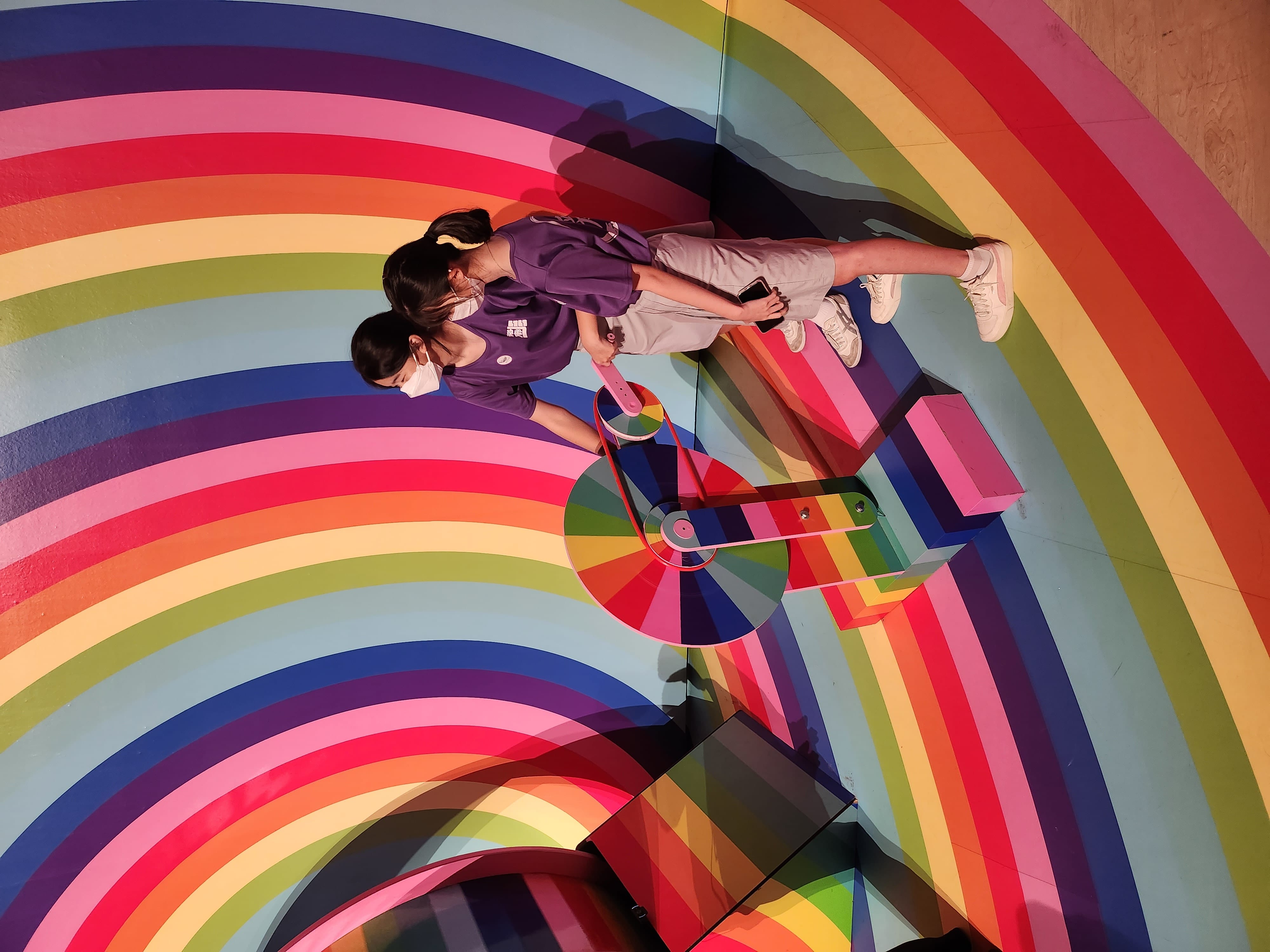 Two masked girls in purple shirts interact with a spinning wheel exhibit surrounded by vibrant rainbow swirl walls and floor.