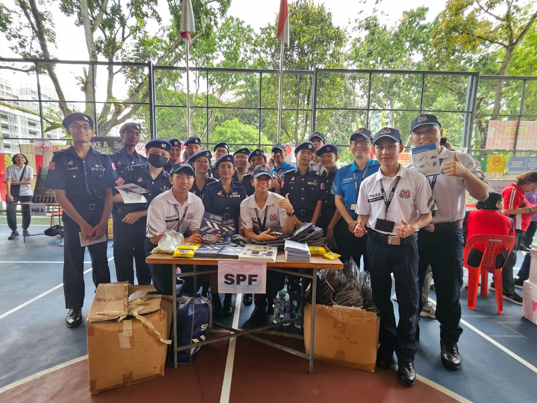 A group of SPF officers and cadets in uniform pose together at an outdoor community event booth with pamphlets.
