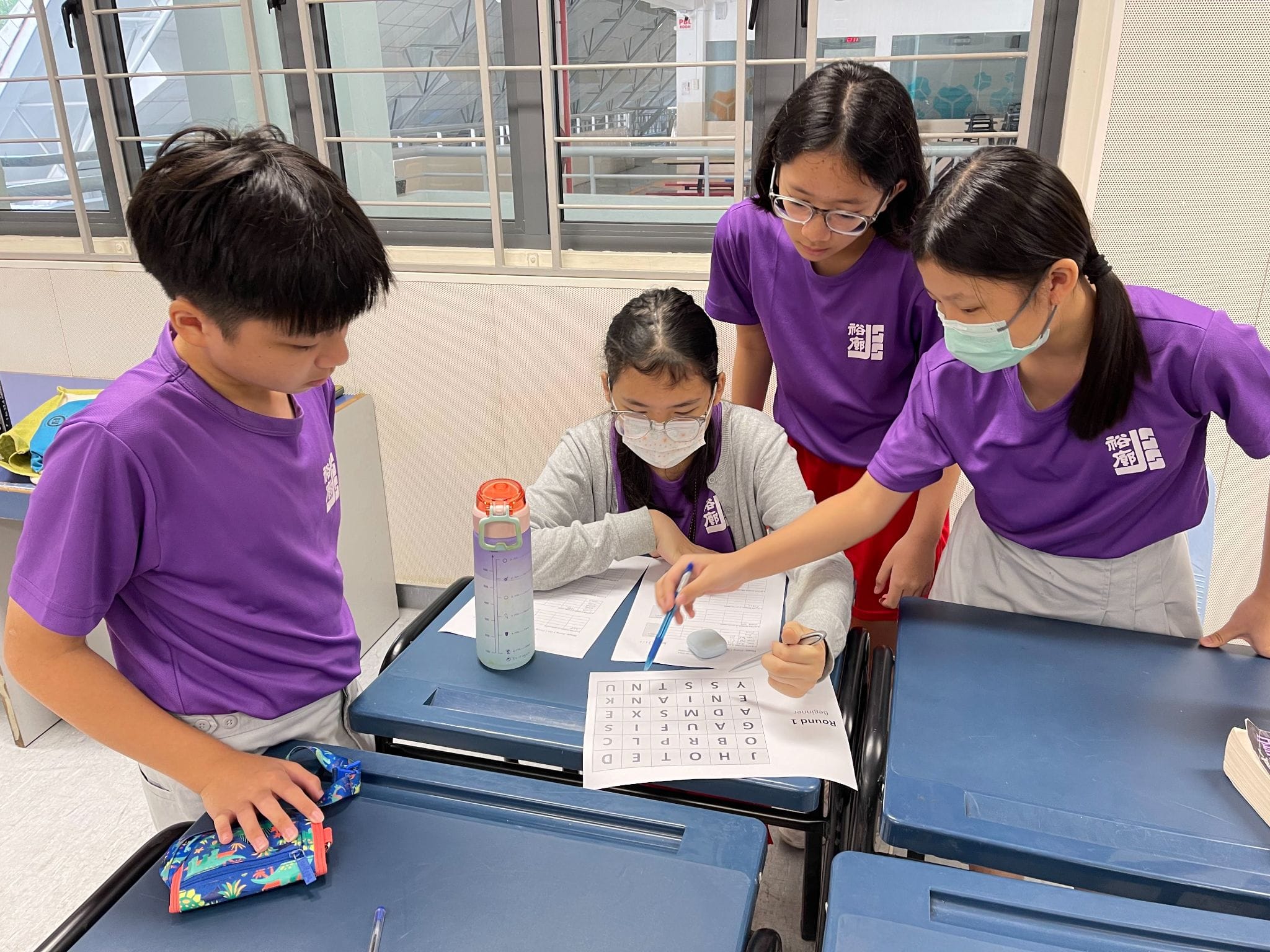 Four students in purple uniforms gather around a desk working on a word search puzzle labeled Round 1.