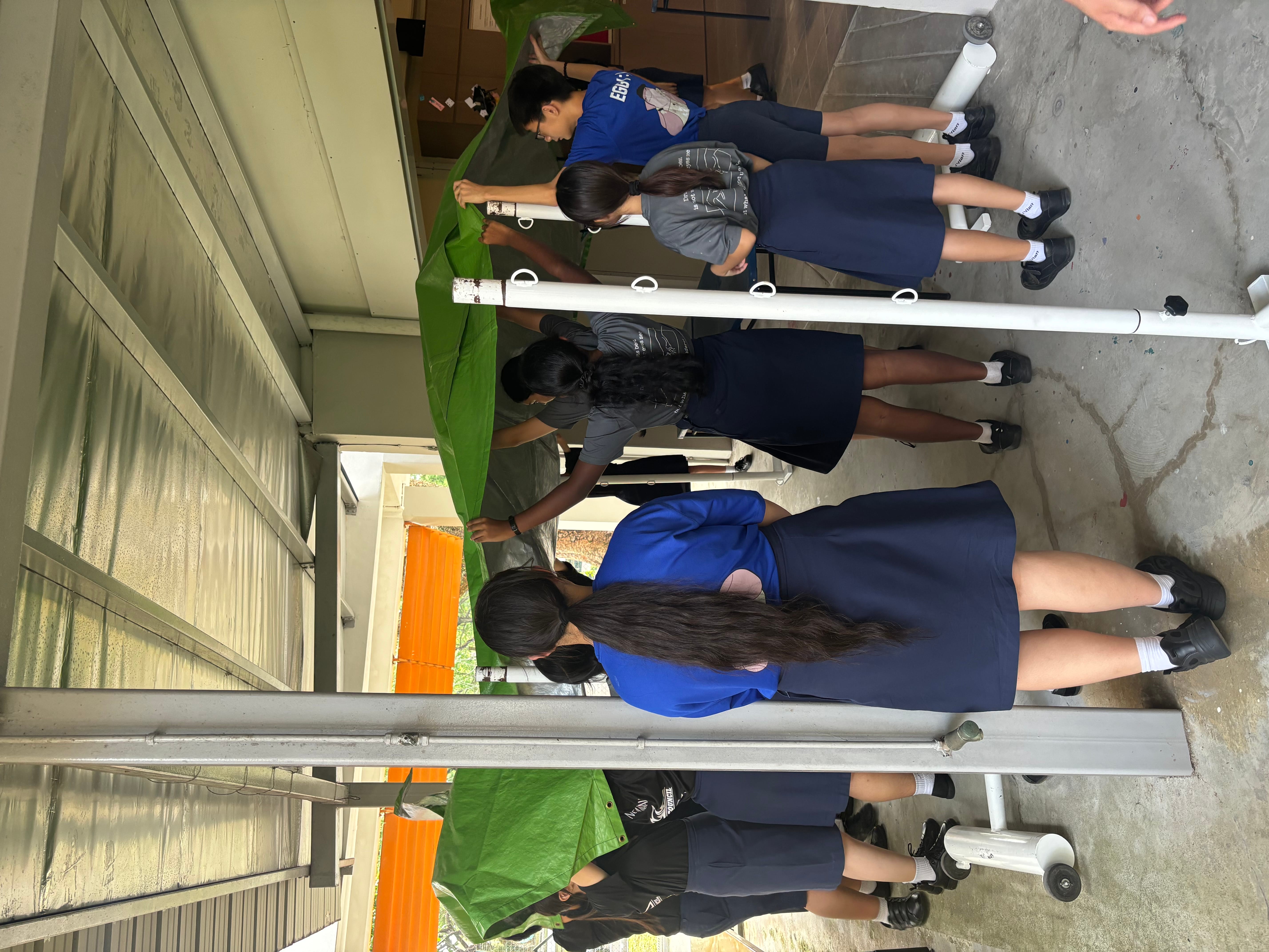 Students in uniforms set up a green tarp canopy on poles in a covered outdoor walkway.