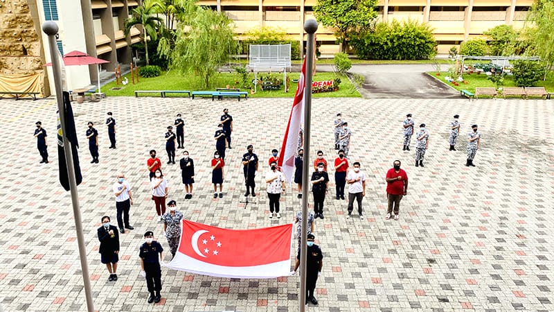 People in uniforms and masks stand spaced apart on a courtyard during a Singapore flag-raising ceremony.