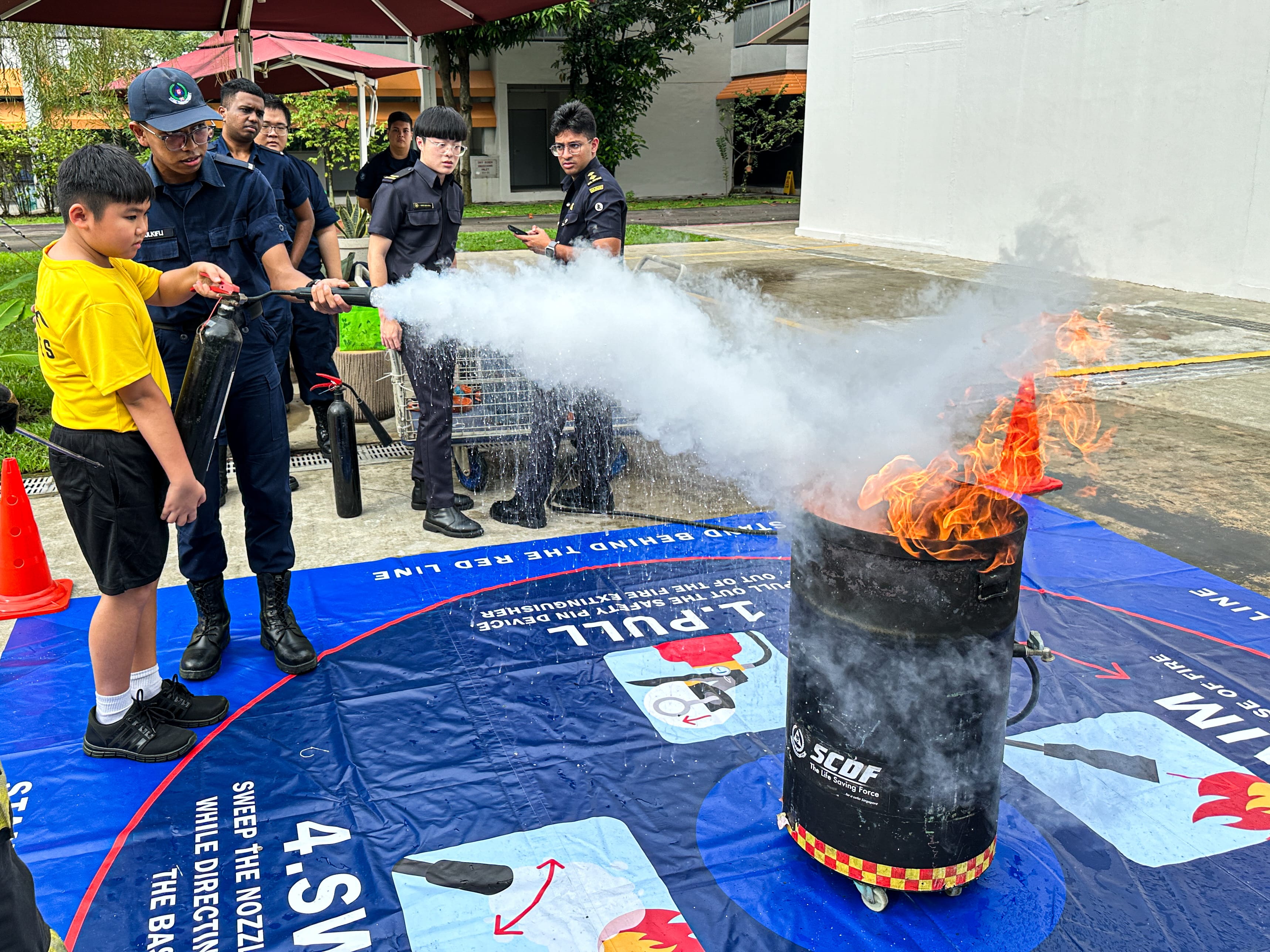 Uniformed instructor helps a boy use a fire extinguisher on a flaming barrel during a safety demo.
