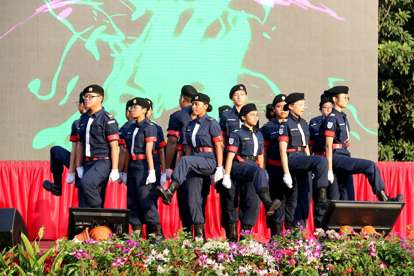 A group of youth in navy blue uniforms with red belts and black berets marching in formation on a stage.