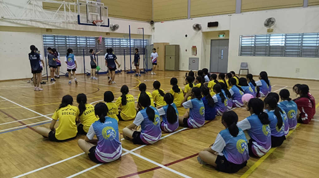 Girls in colorful jerseys sit on an indoor basketball court floor watching a small group practice a sport nearby.