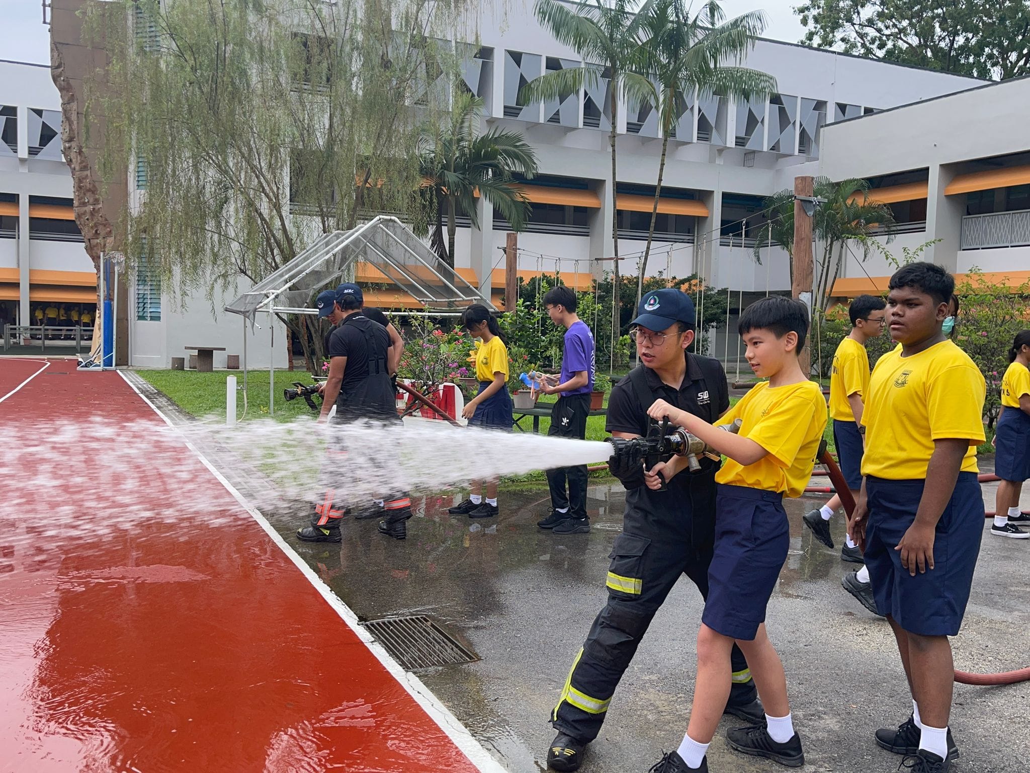 A student in yellow aims a fire hose with an instructor's help at a school fire drill, while other students watch.