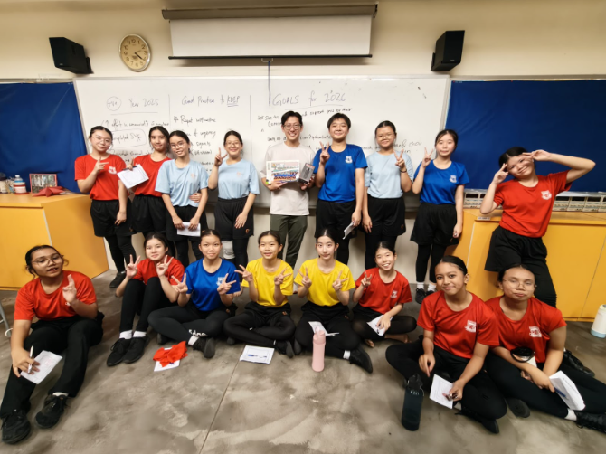 Group of students in red, blue, and yellow shirts posing with peace signs in front of a whiteboard in a classroom.
