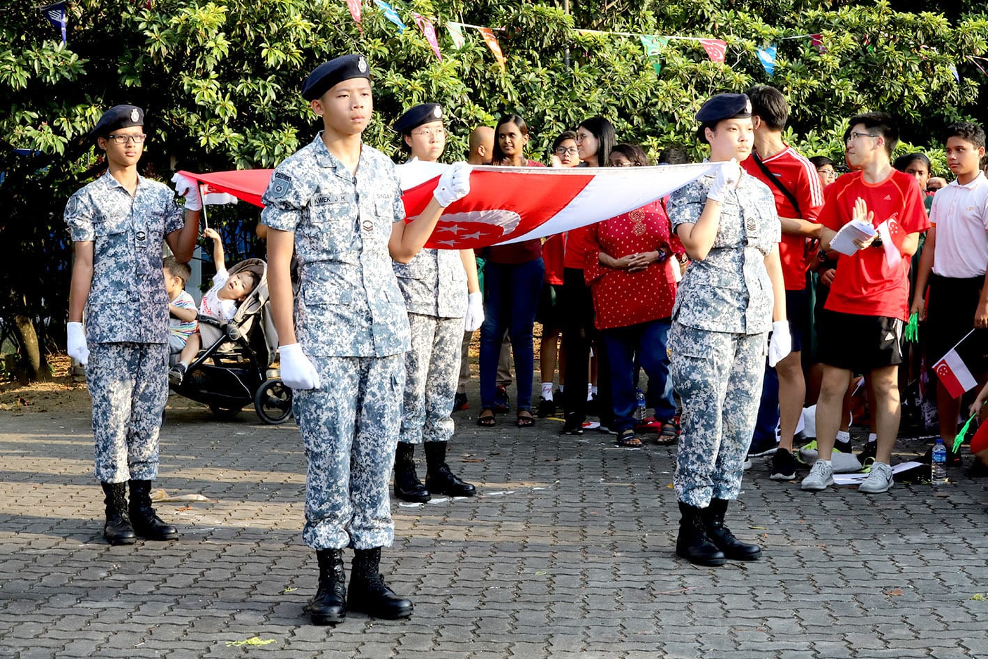 Young cadets in camouflage uniforms and berets carry the Singapore flag during an outdoor ceremony with a crowd watching.