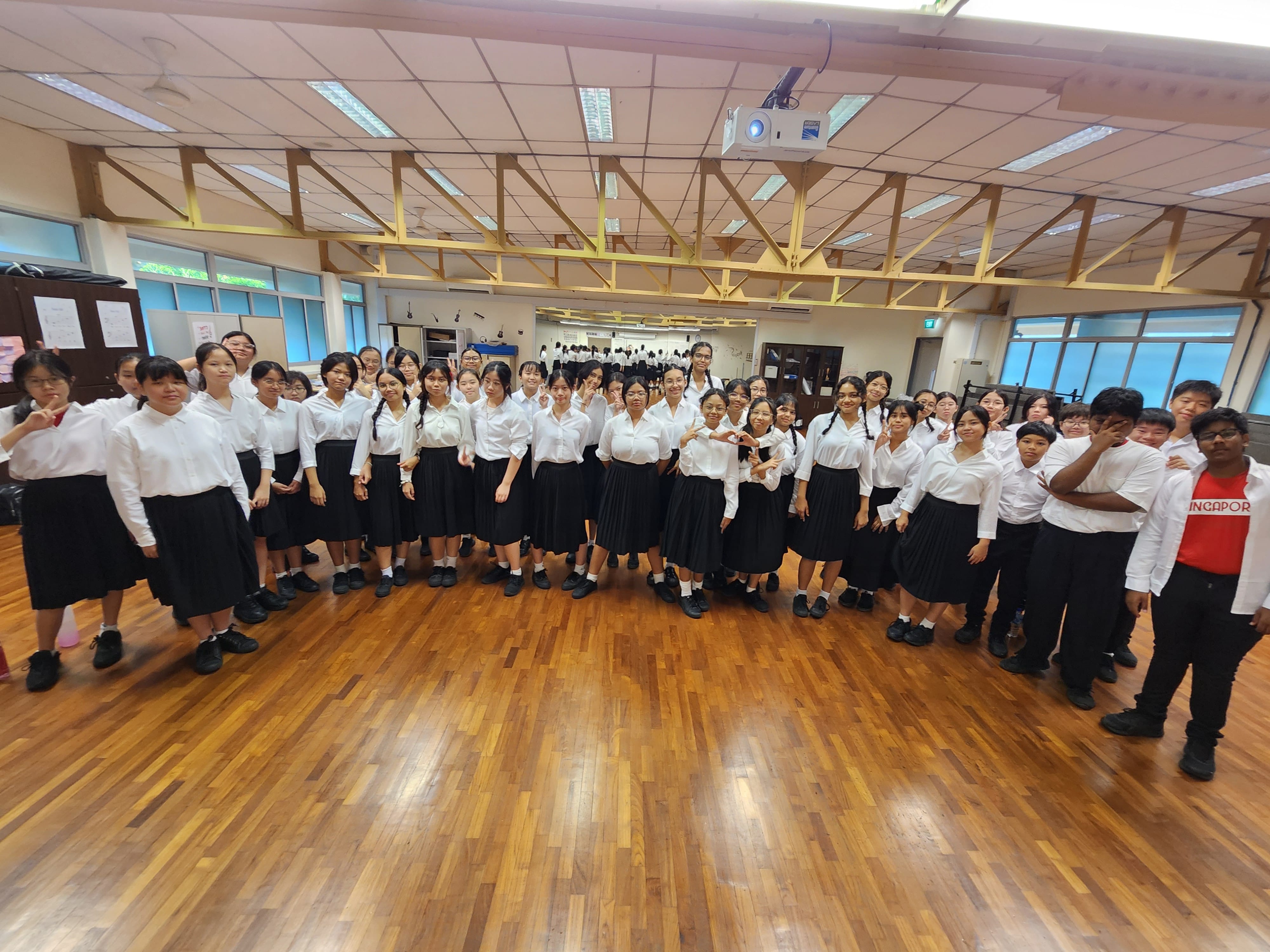 A large group of students in white shirts and black skirts pose together in a school hall with wooden floors.