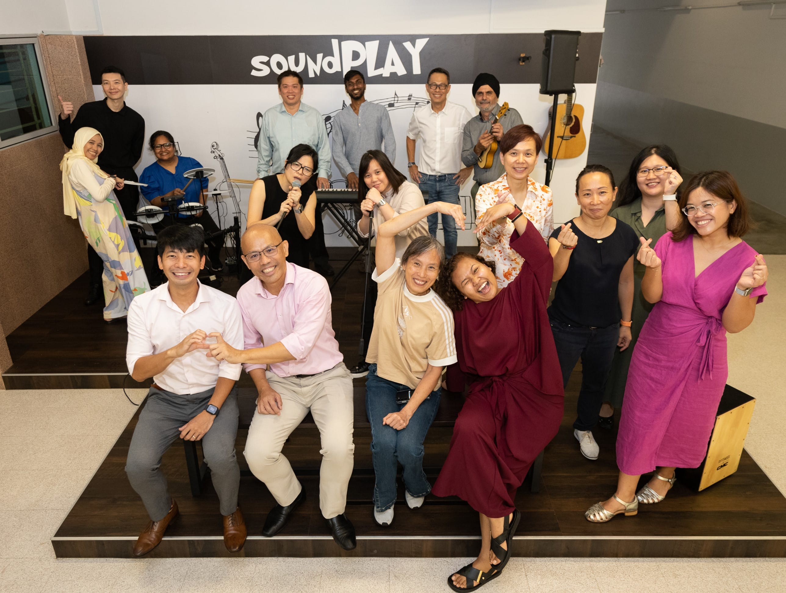 A large group of smiling people pose together in front of a wall with the text SoundPLAY, with musical instruments nearby.