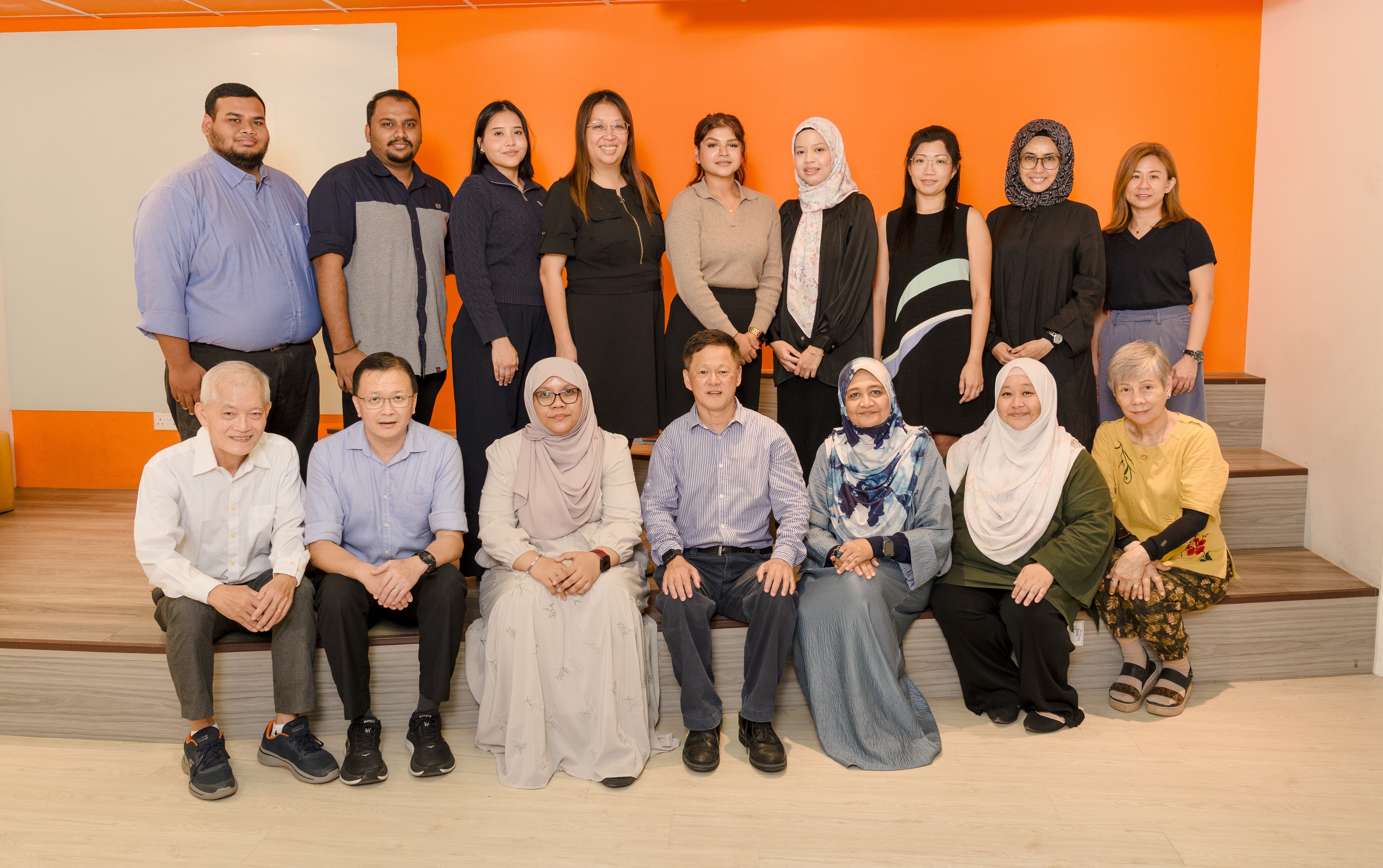 A group of about 15 people posed in two rows indoors against an orange wall, some seated and some standing.