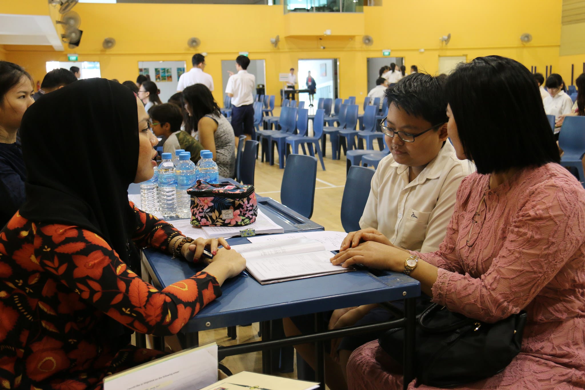 Three people discuss documents at a blue table in a busy school hall with yellow walls and blue chairs.