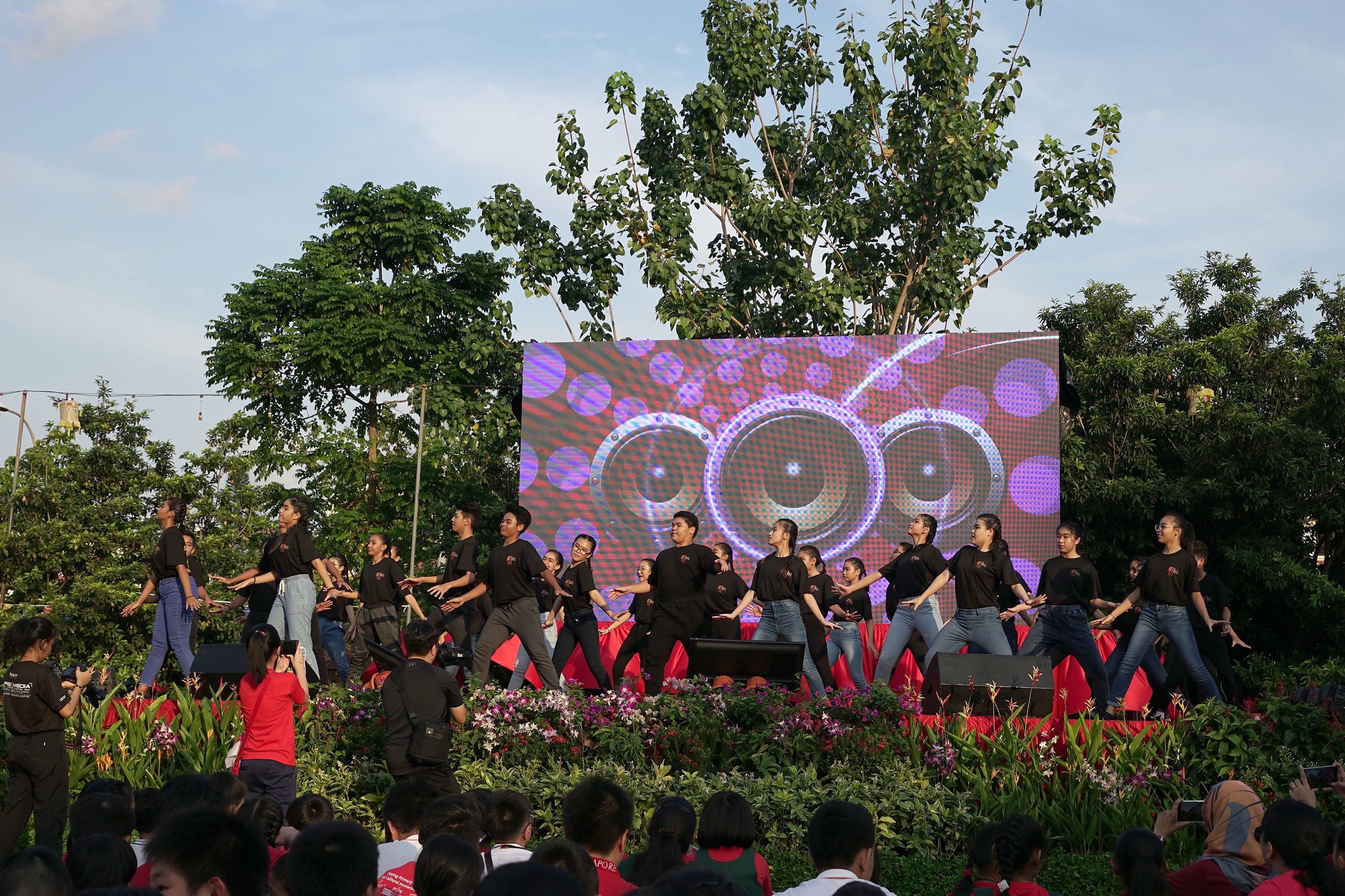 A group of dancers in black shirts perform on an outdoor stage with a colorful LED screen backdrop and crowd watching.