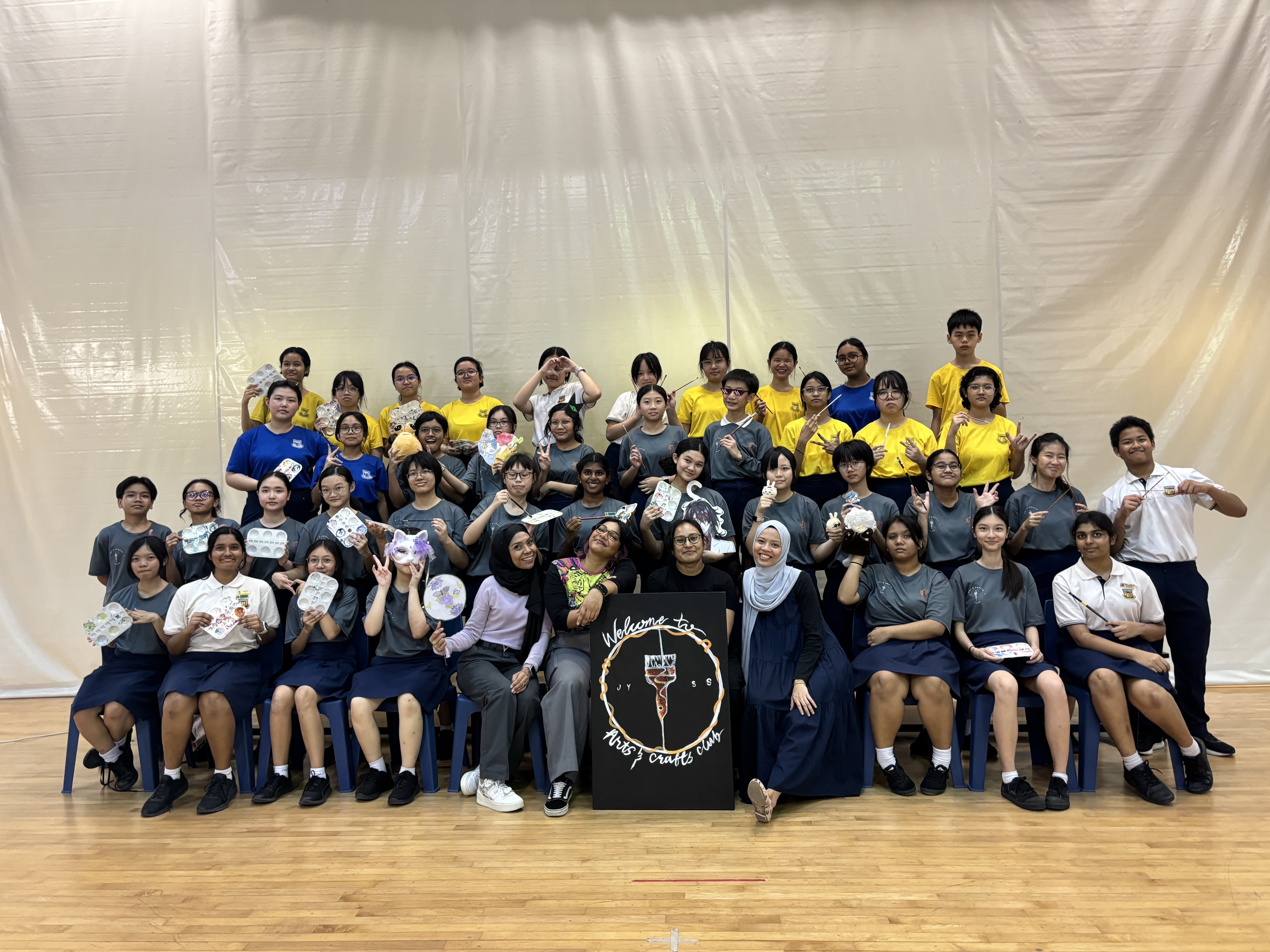 A large group of students pose with crafts and a black sign reading "Welcome to Arts and Crafts Club."