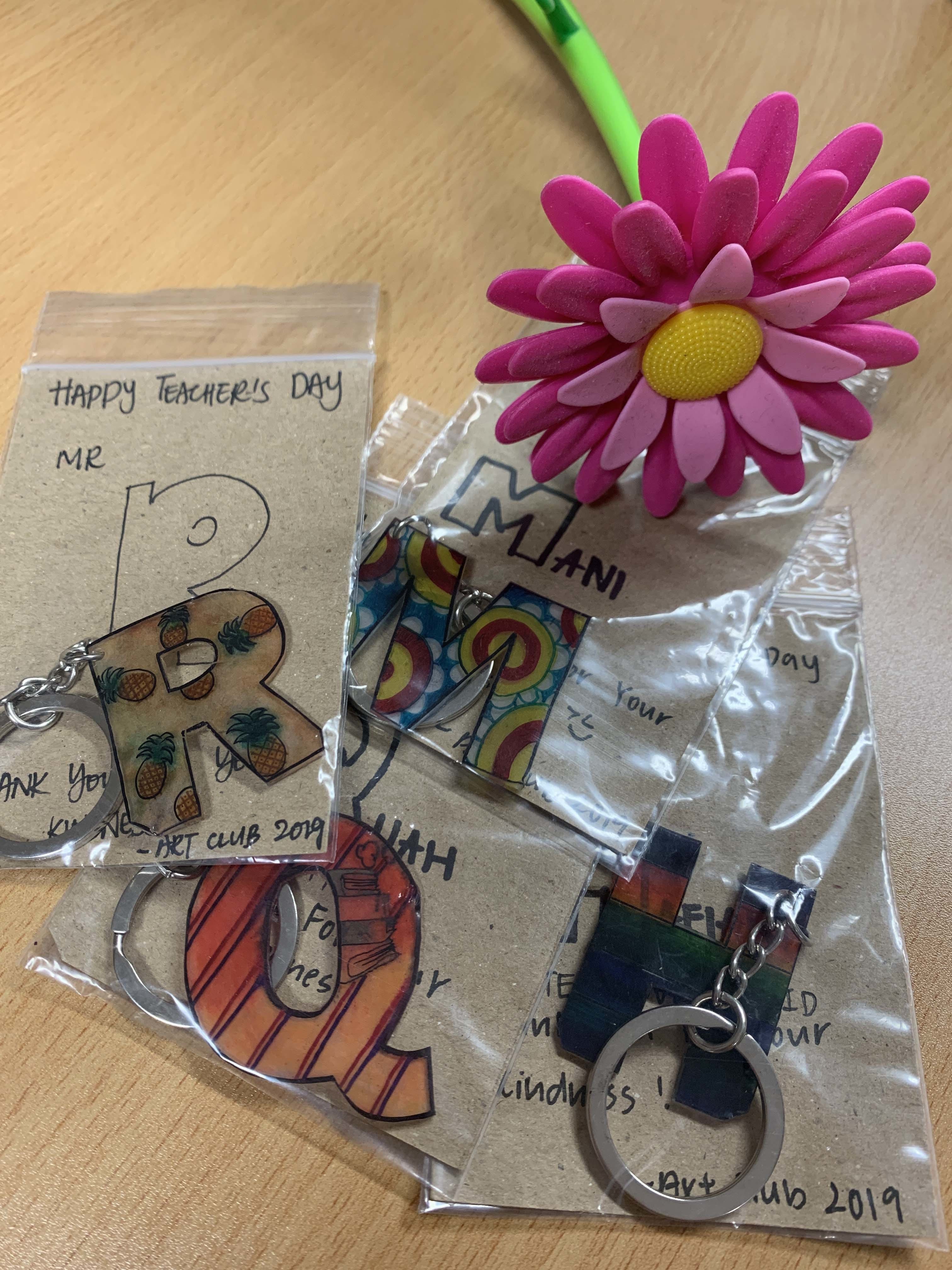 Colorful letter-shaped keychains in gift bags labeled Happy Teachers Day from Art Club 2019, beside a pink flower pen.