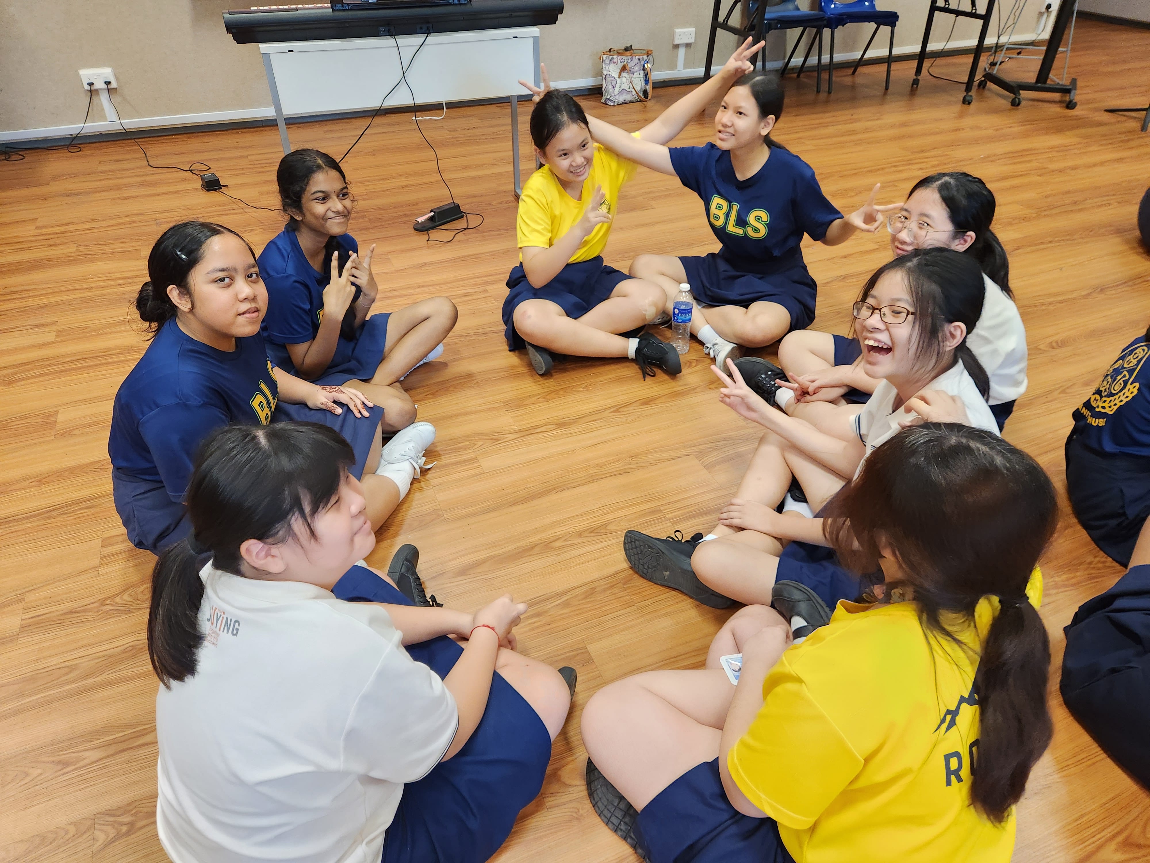 A group of school girls in blue and yellow uniforms sit in a circle on a wooden floor, smiling and making peace signs.