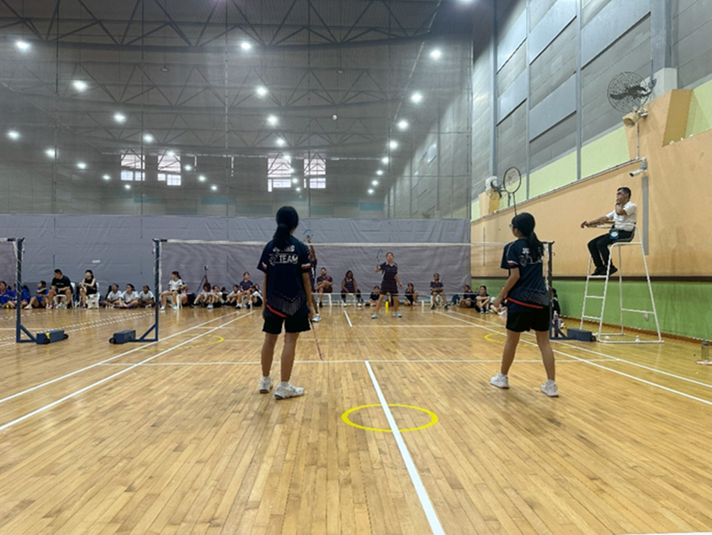 Two players in dark uniforms play badminton indoors with a referee on a chair and spectators seated along the wall.