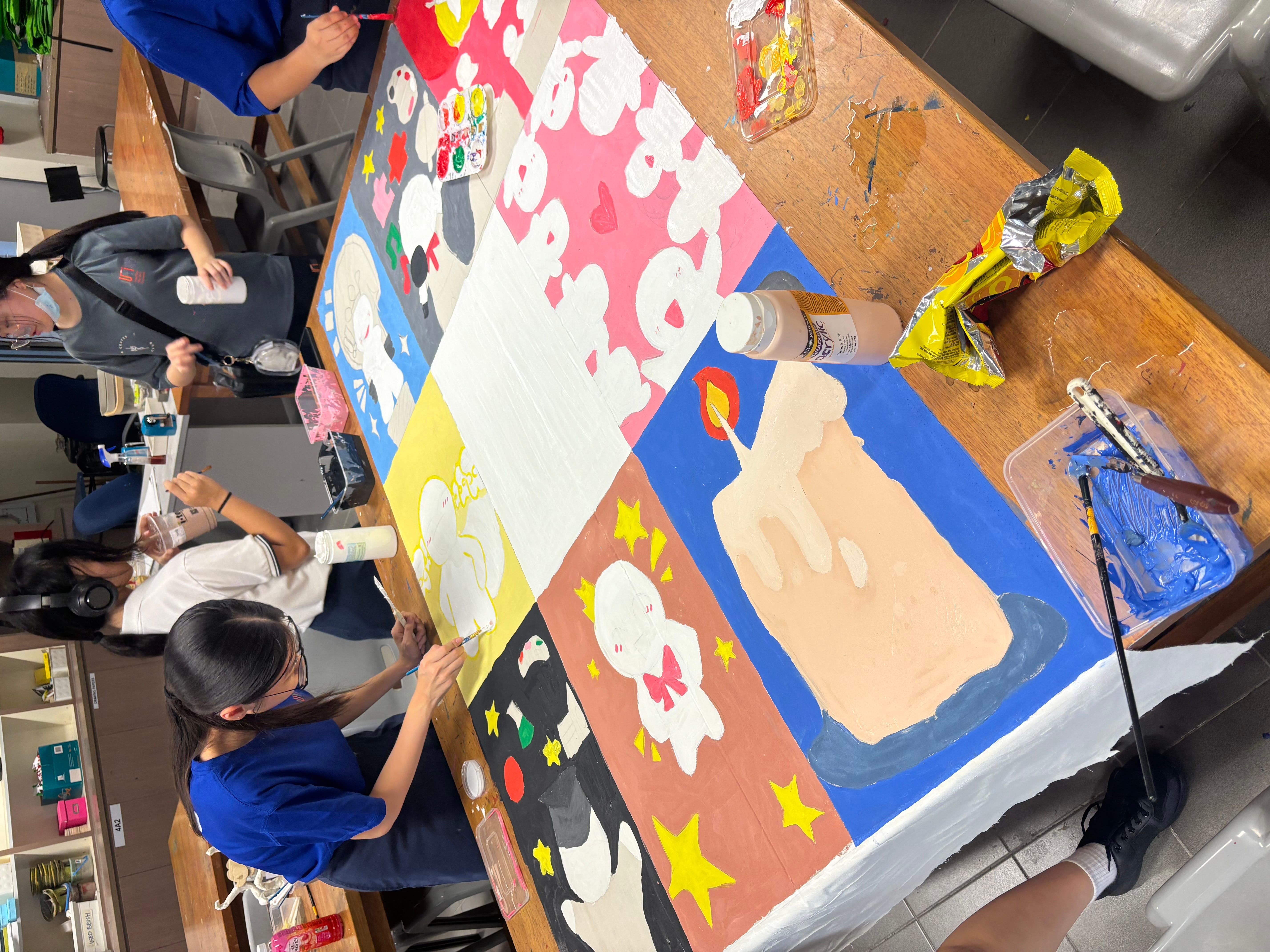 Students paint a colorful mural on a long table, with paint trays, brushes, and a glue bottle nearby.