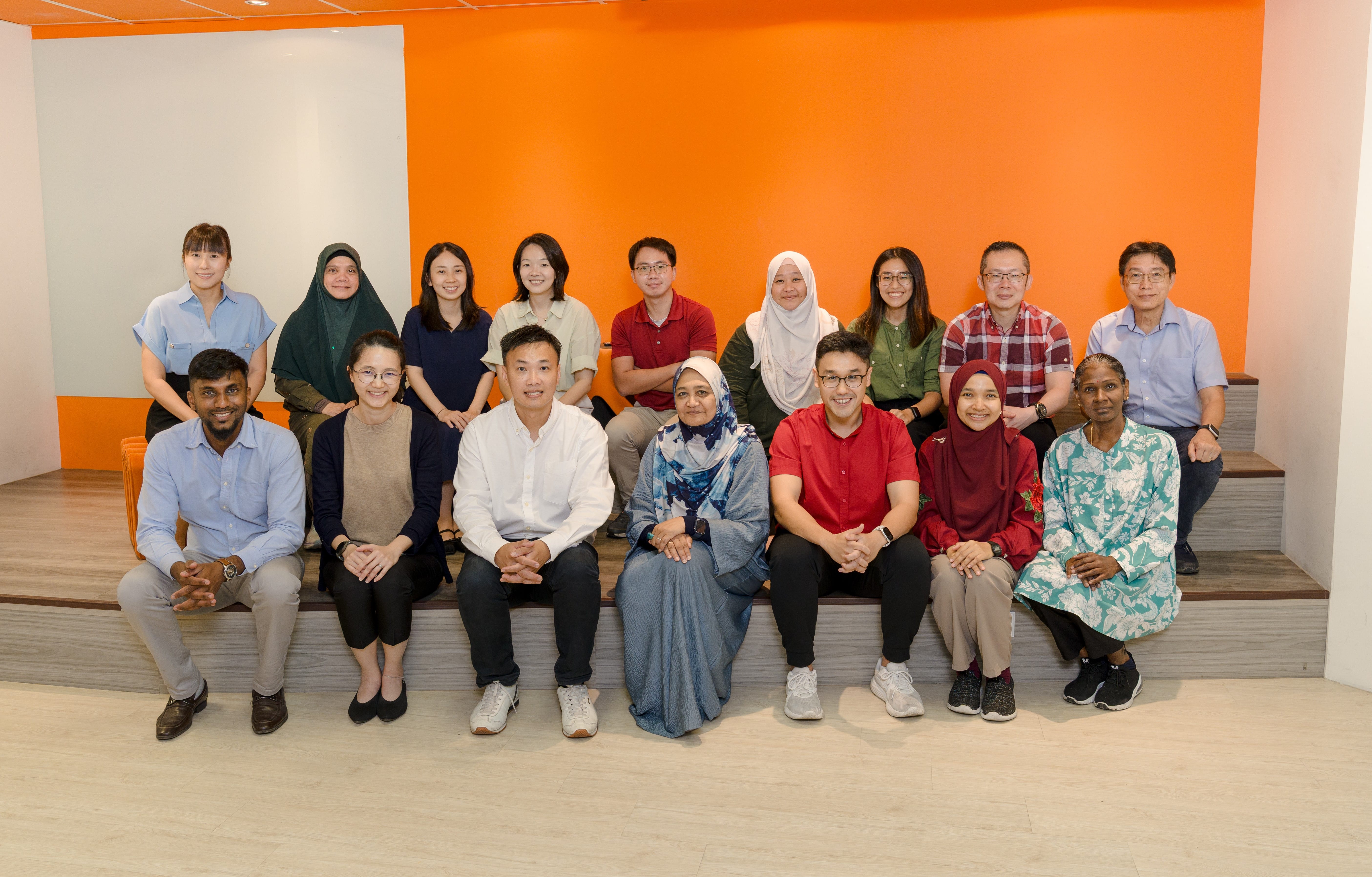 A diverse group of about 16 people posing together in two rows against a bright orange wall indoors.