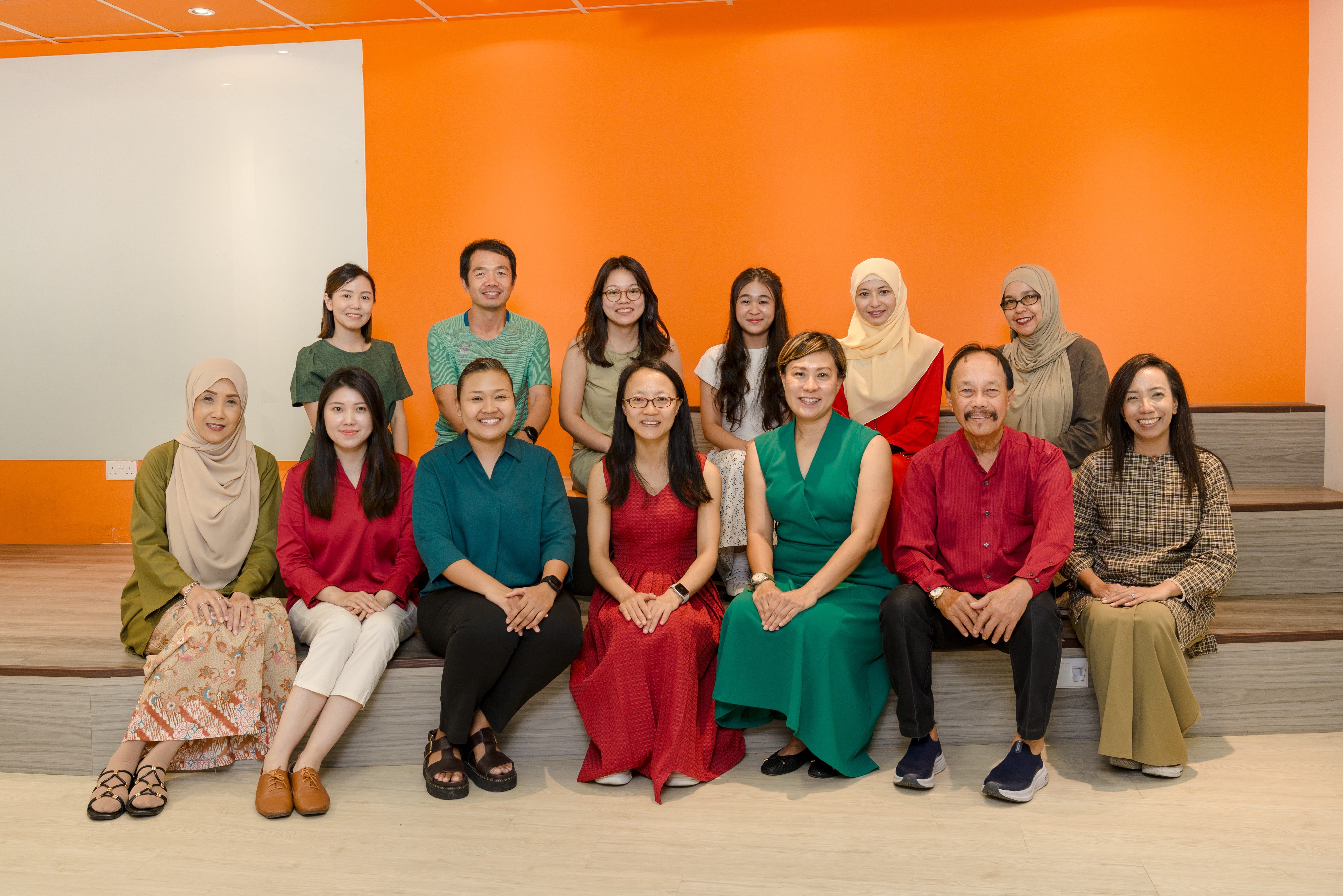 Group portrait of 12 people seated on tiered benches against a bright orange wall.