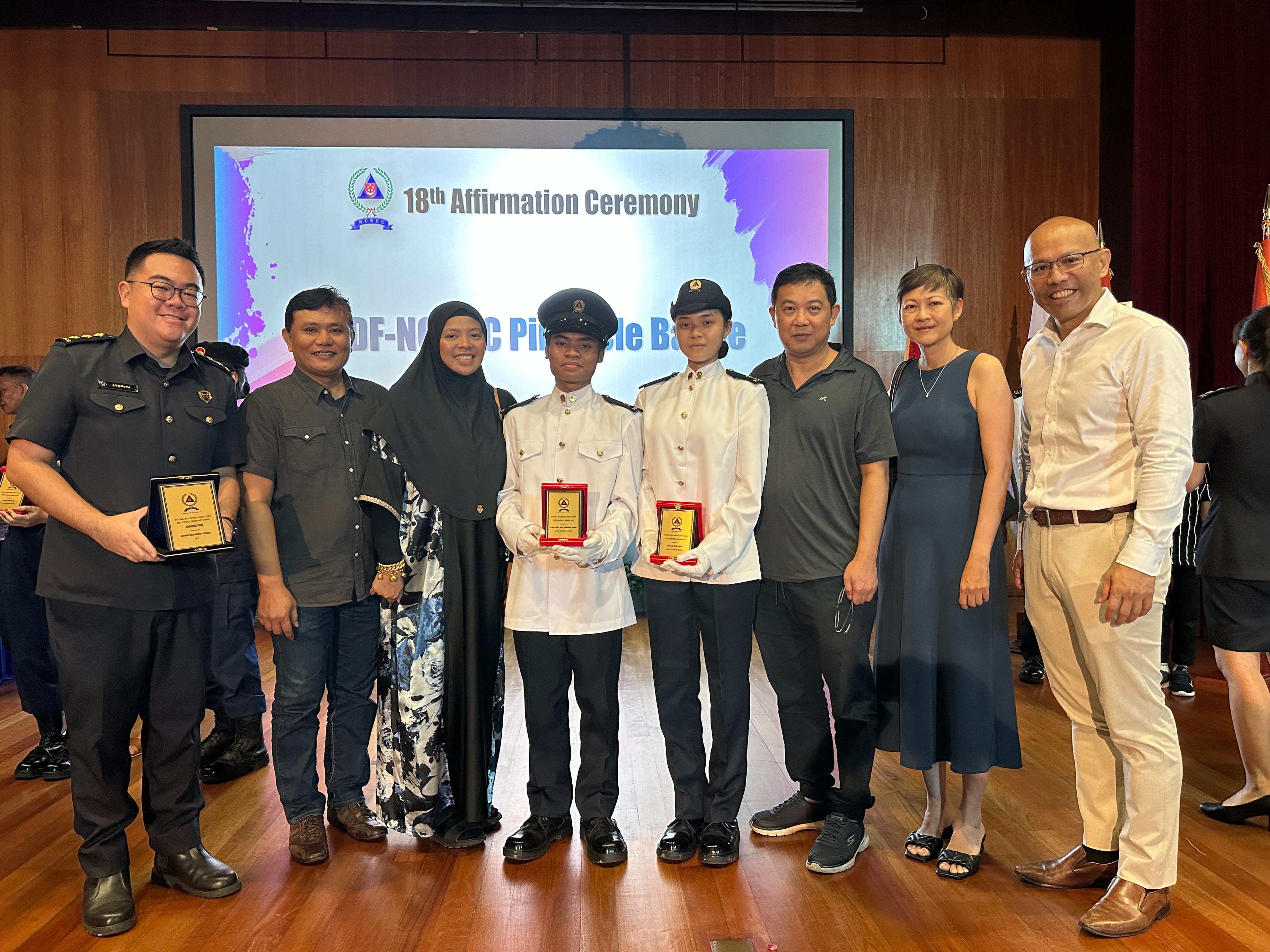 Eight people pose at the 18th Affirmation Ceremony; two uniformed cadets hold award plaques in the center.