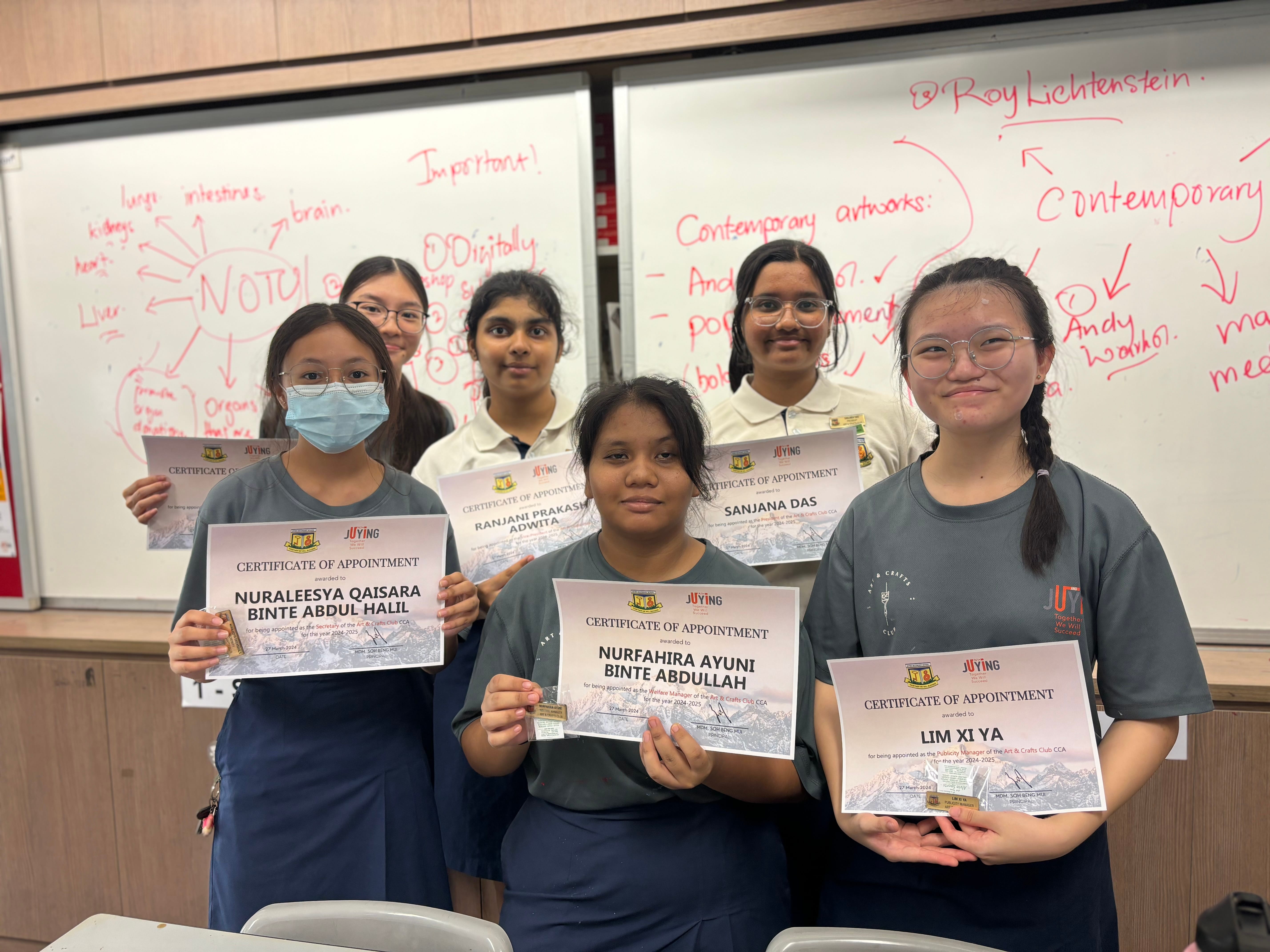 Six female students hold certificates of appointment in a classroom with a whiteboard covered in notes.