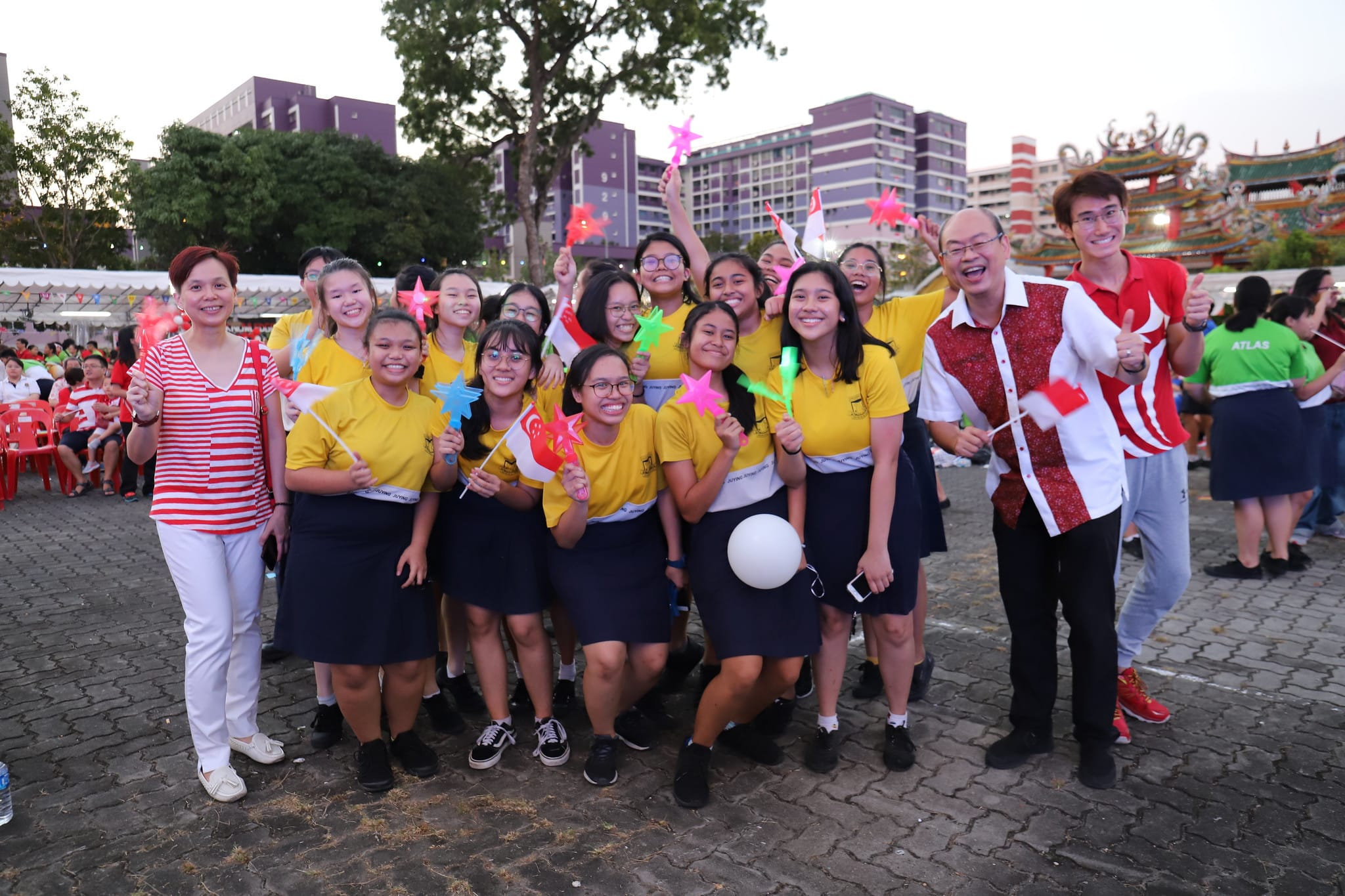 A cheerful group of students in yellow uniforms holds small flags and colorful star wands at an outdoor celebration.