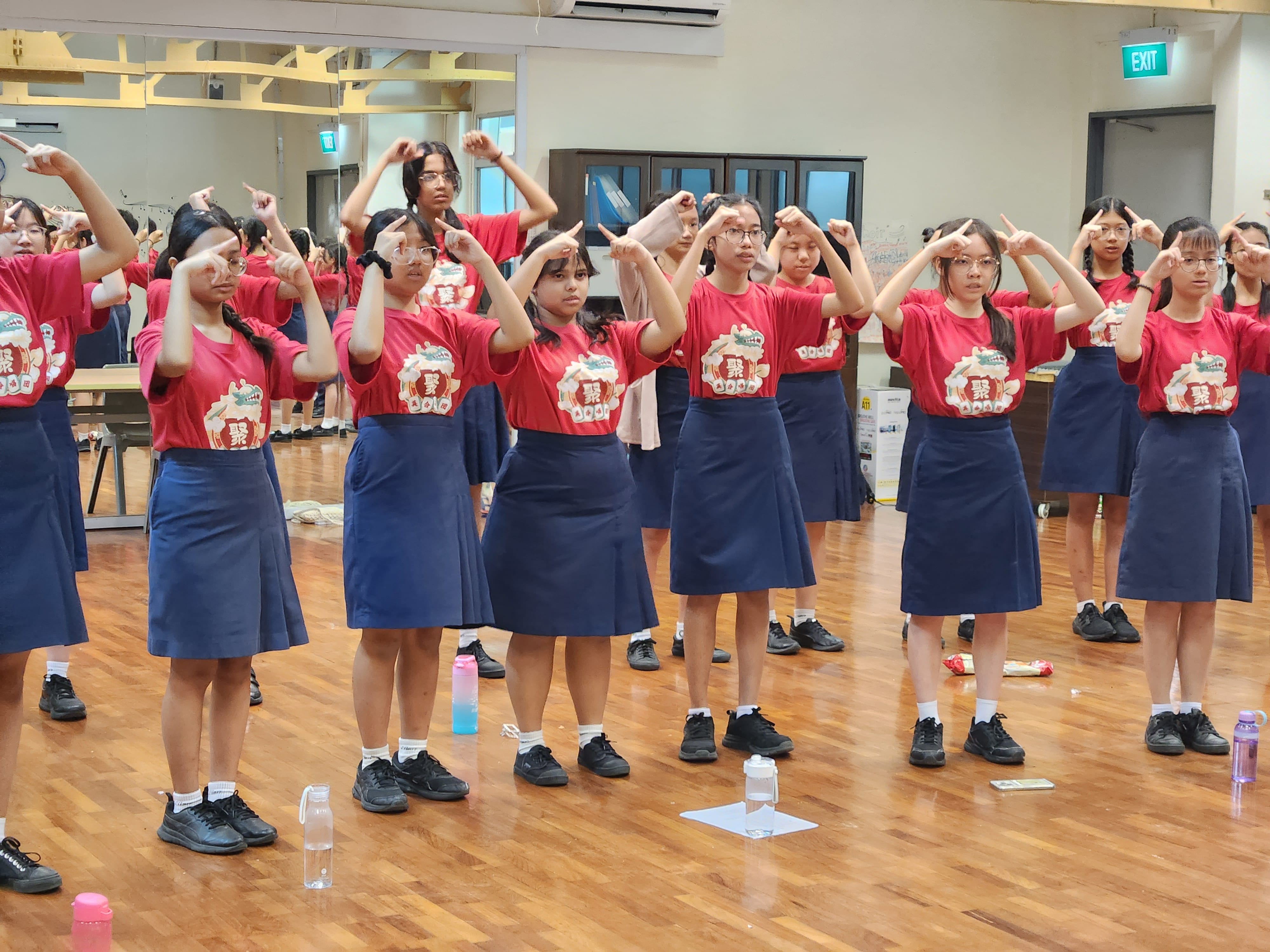 Girls in red shirts and blue skirts stand in rows on a wooden floor, raising their hands above their heads in unison.