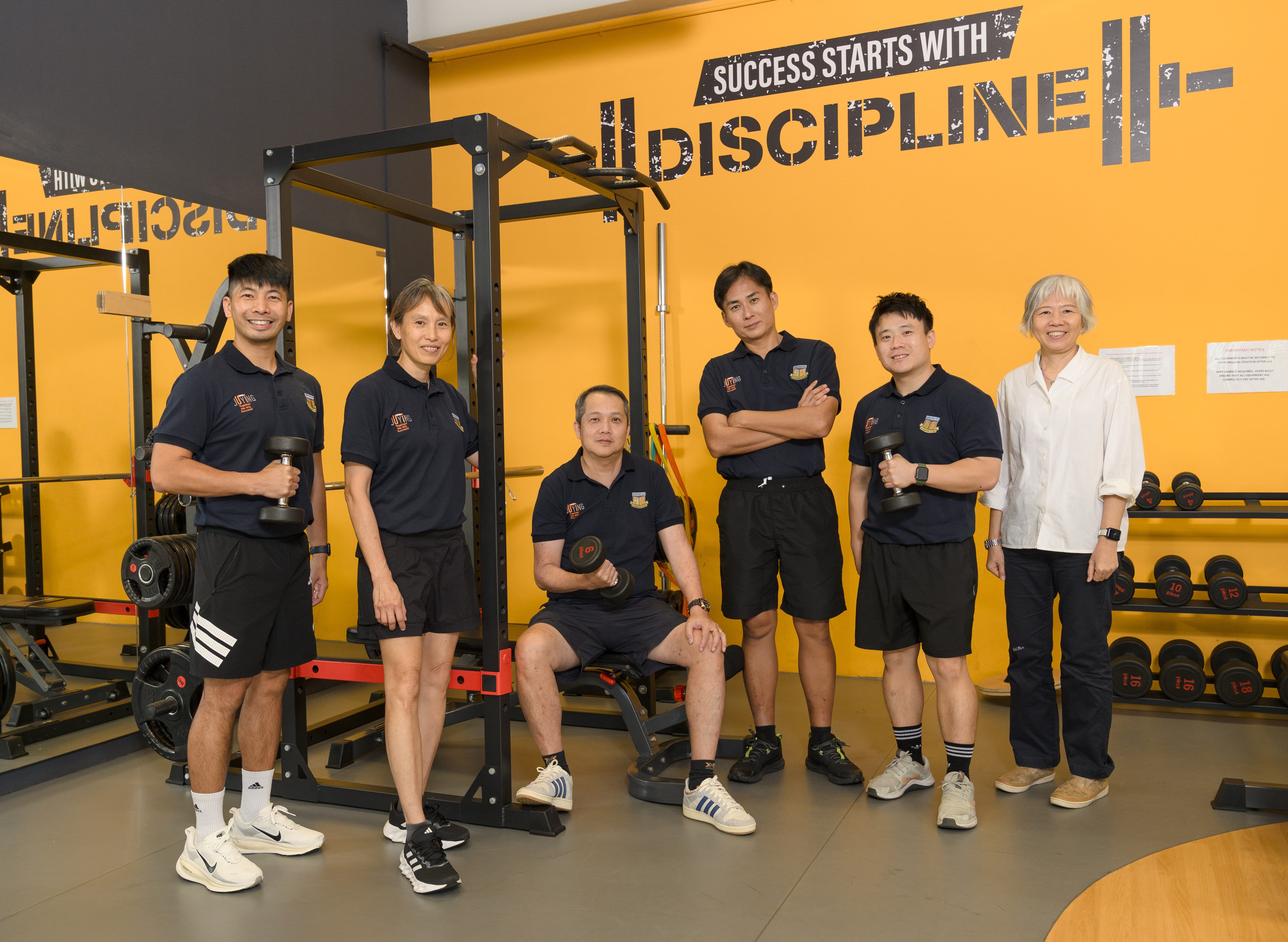 Six people pose in a gym by a squat rack and dumbbells, with a wall sign reading SUCCESS STARTS WITH DISCIPLINE.