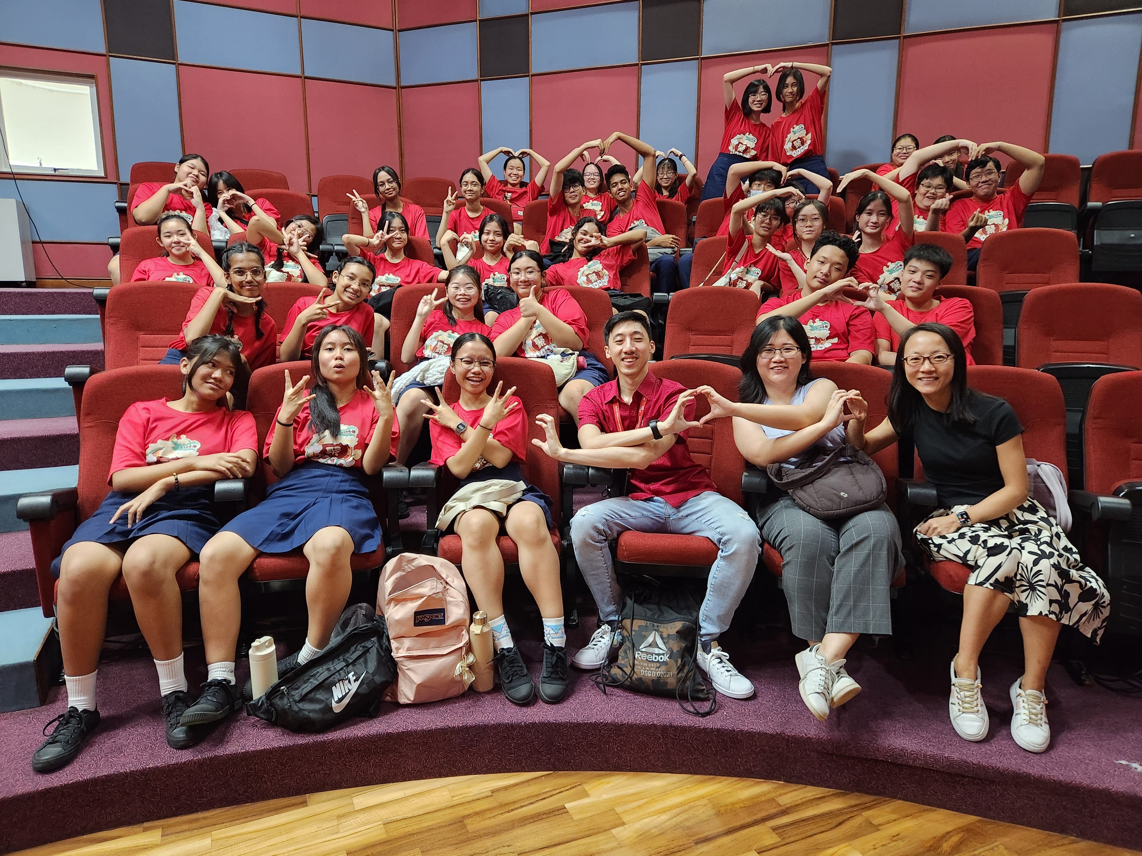 Large group of students in red shirts sitting in red auditorium seats, smiling and making heart hand gestures.