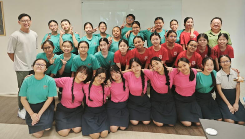 A large group of students in teal, red, and pink shirts pose together indoors, many making peace signs.