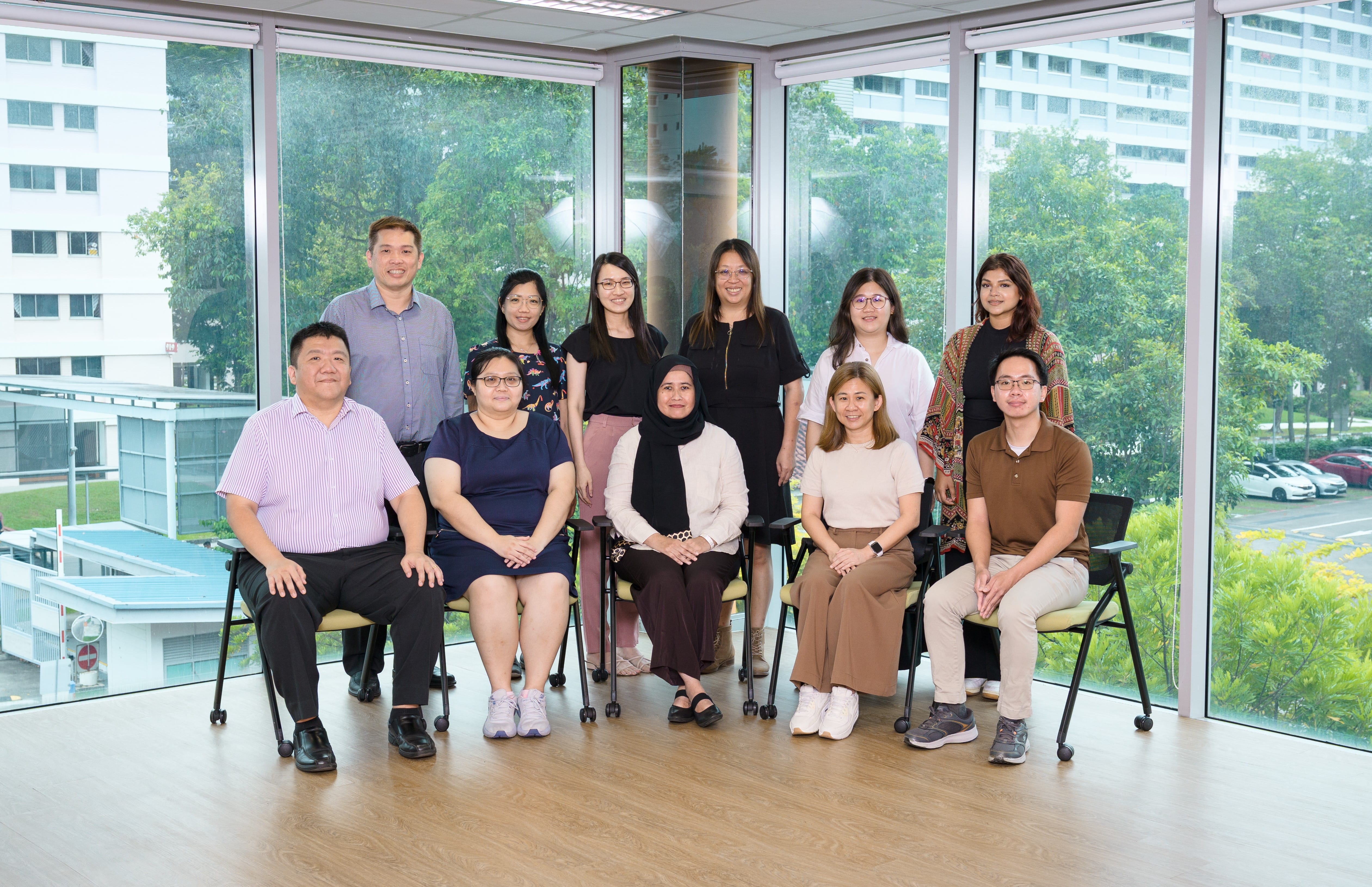 Eleven people pose for a group photo in an office with large windows overlooking trees and buildings.