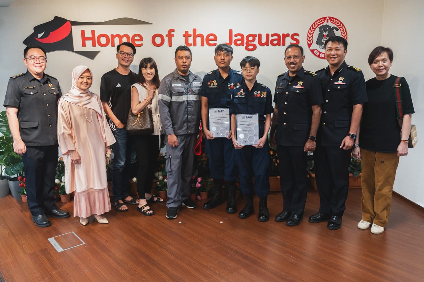 Ten people pose indoors before a wall reading Home of the Jaguars; two in uniform hold SCDF award certificates.