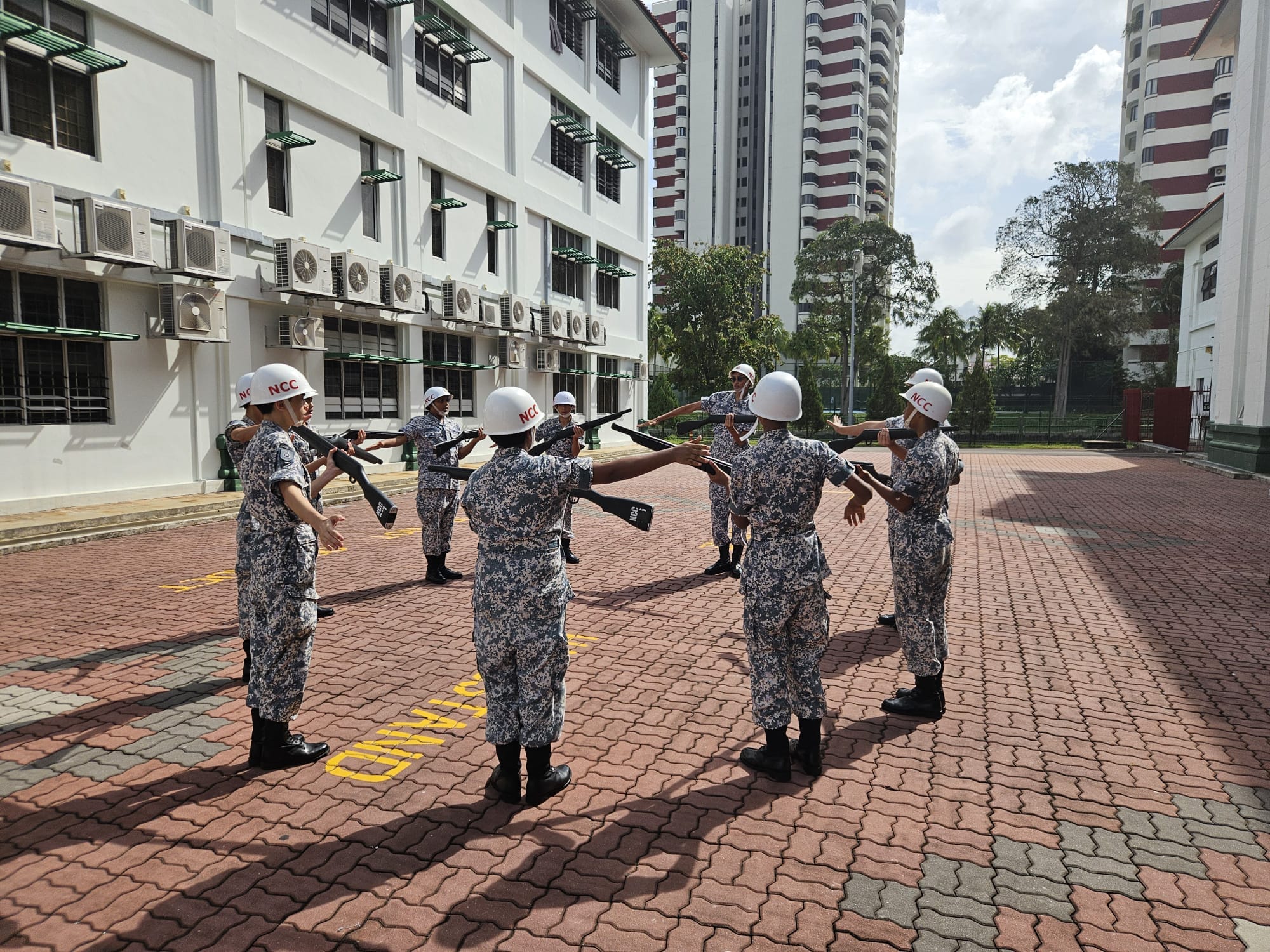 NCC cadets in camouflage uniforms and white helmets practice rifle drill on a brick courtyard beside white buildings.